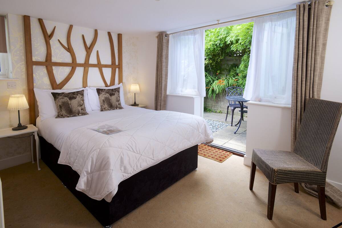 A bedroom with a large bed, a chair, a nightstand with a lamp, and a view of the garden through the window.