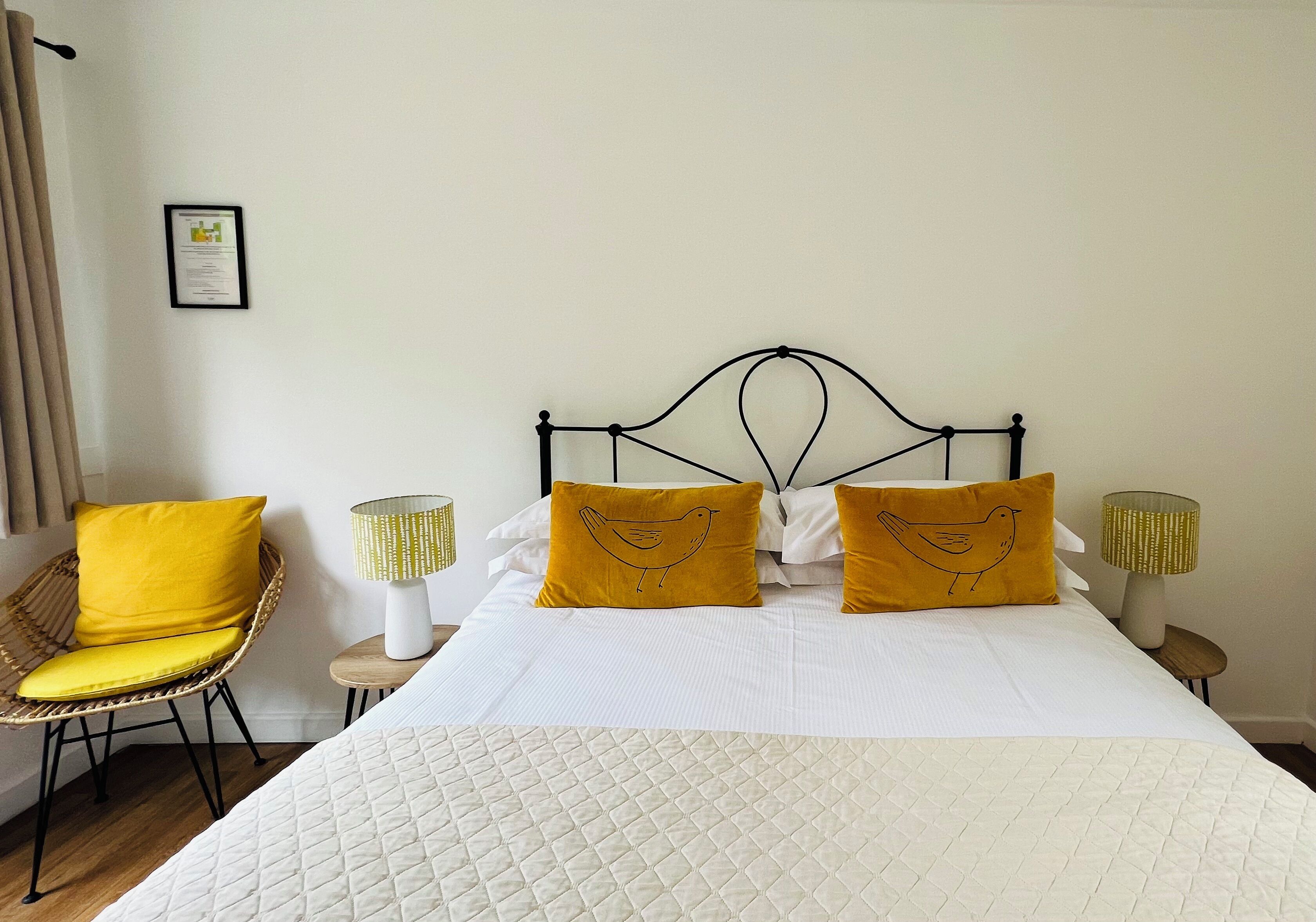 Un lit bien fait, avec une literie blanche et deux oreillers jaunes, une chaise en osier avec un coussin jaune, et deux lampes de chevet avec des abat-jour jaunes.