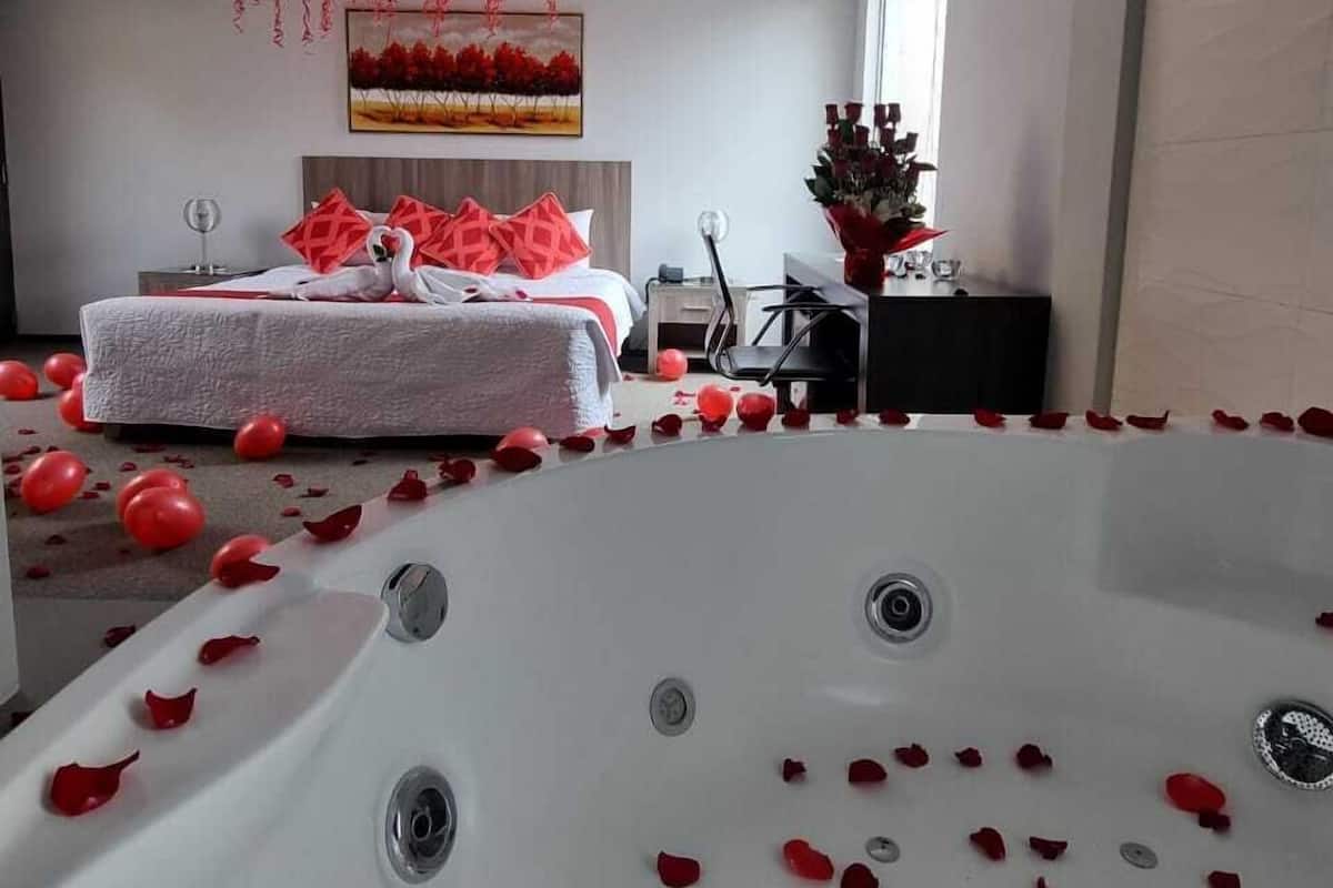 A modern bedroom with a large bathtub, red balloons, and rose petals scattered on the floor.