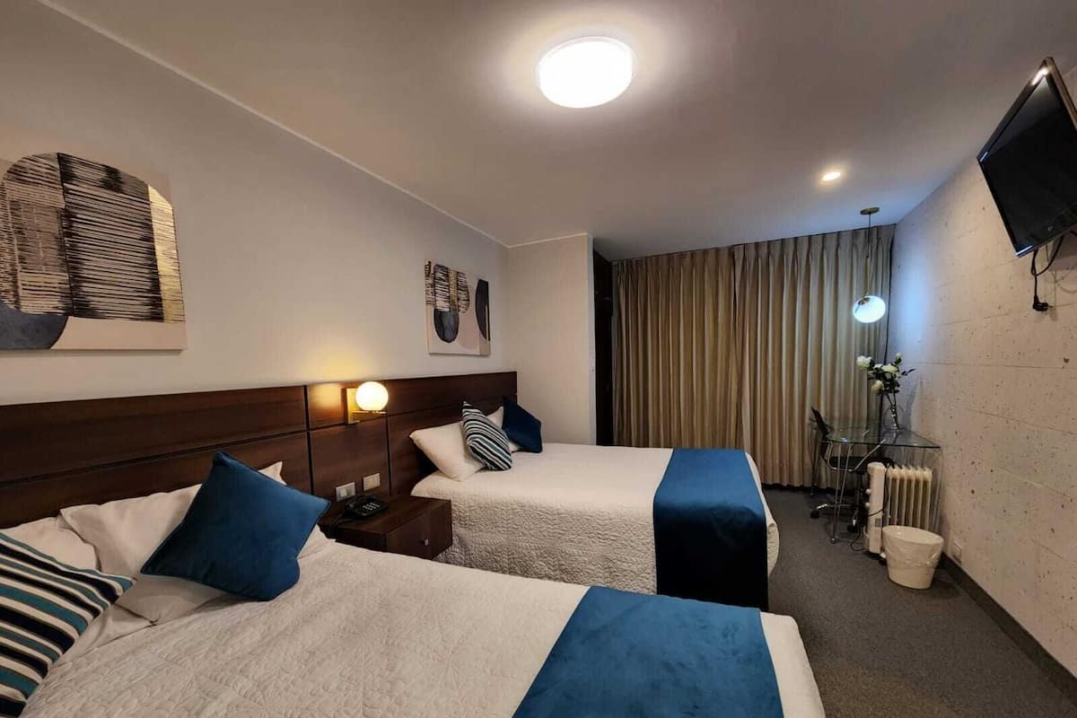 A hotel room with two beds, a desk with a lamp, a TV, and a radiator.
