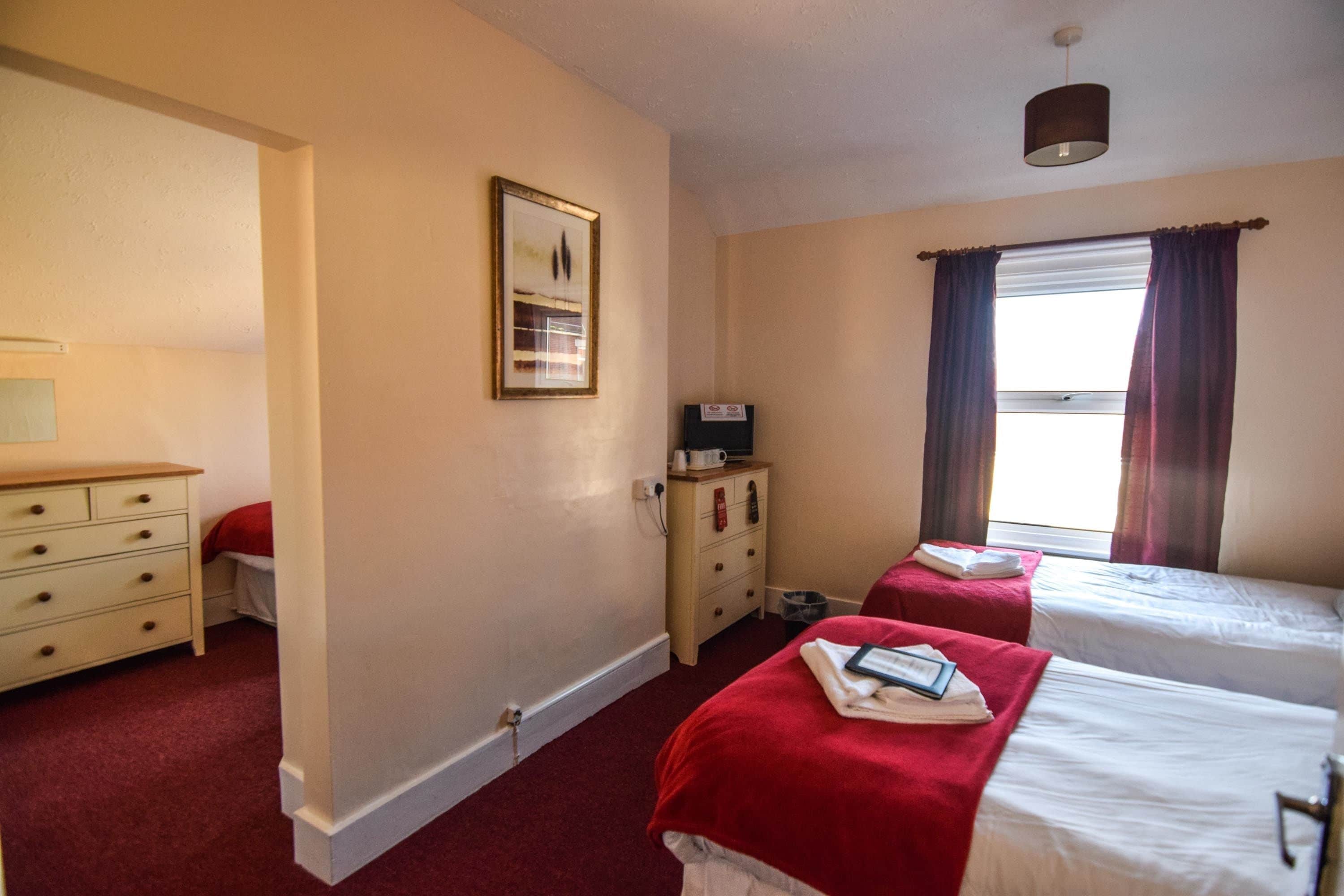A hotel room with two single beds, a wooden dresser, a nightstand, a framed picture on the wall, and a window with curtains.