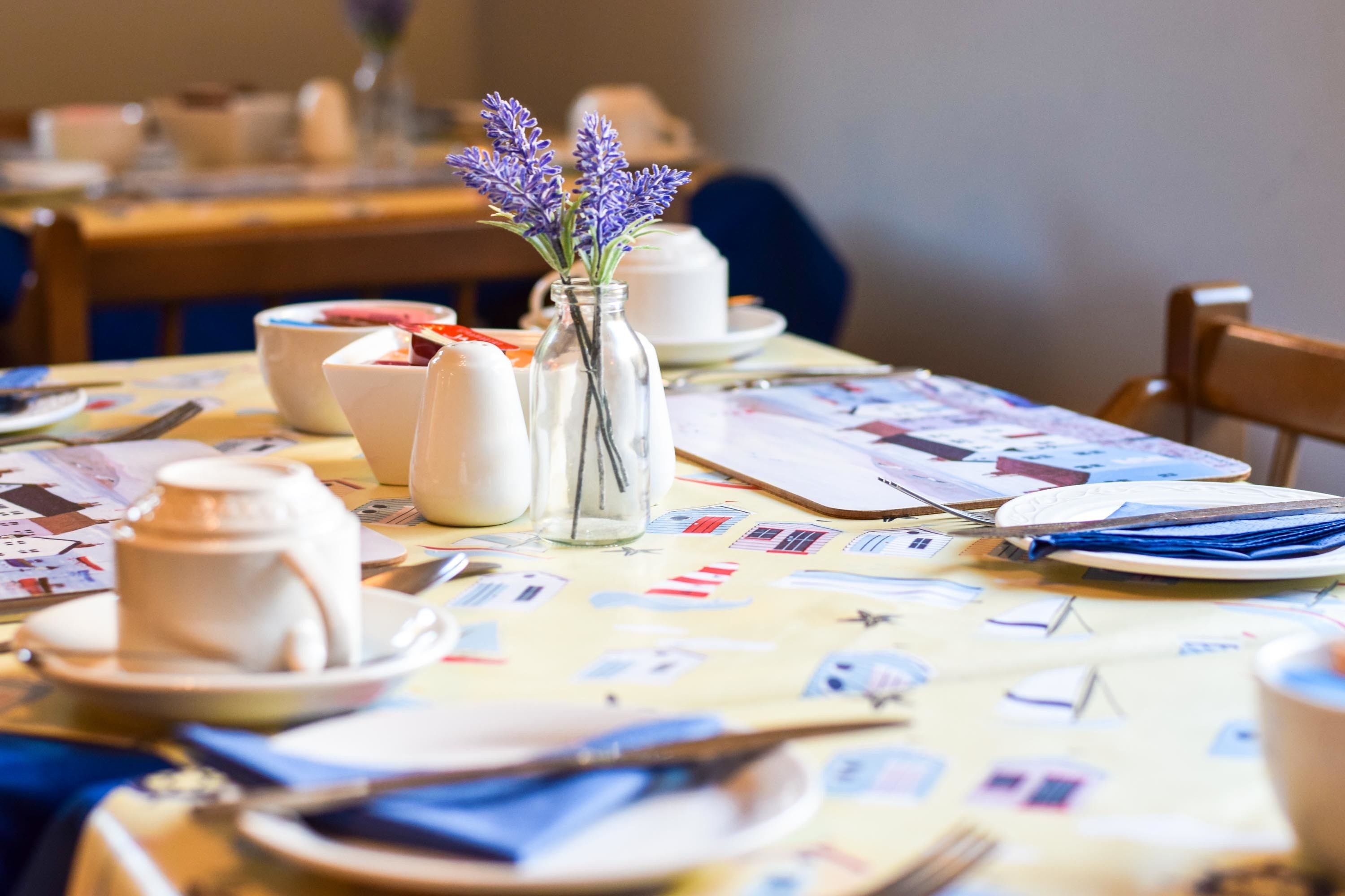 A dining table set with white ceramic dishes, a vase with purple flowers, and a colorful tablecloth.
