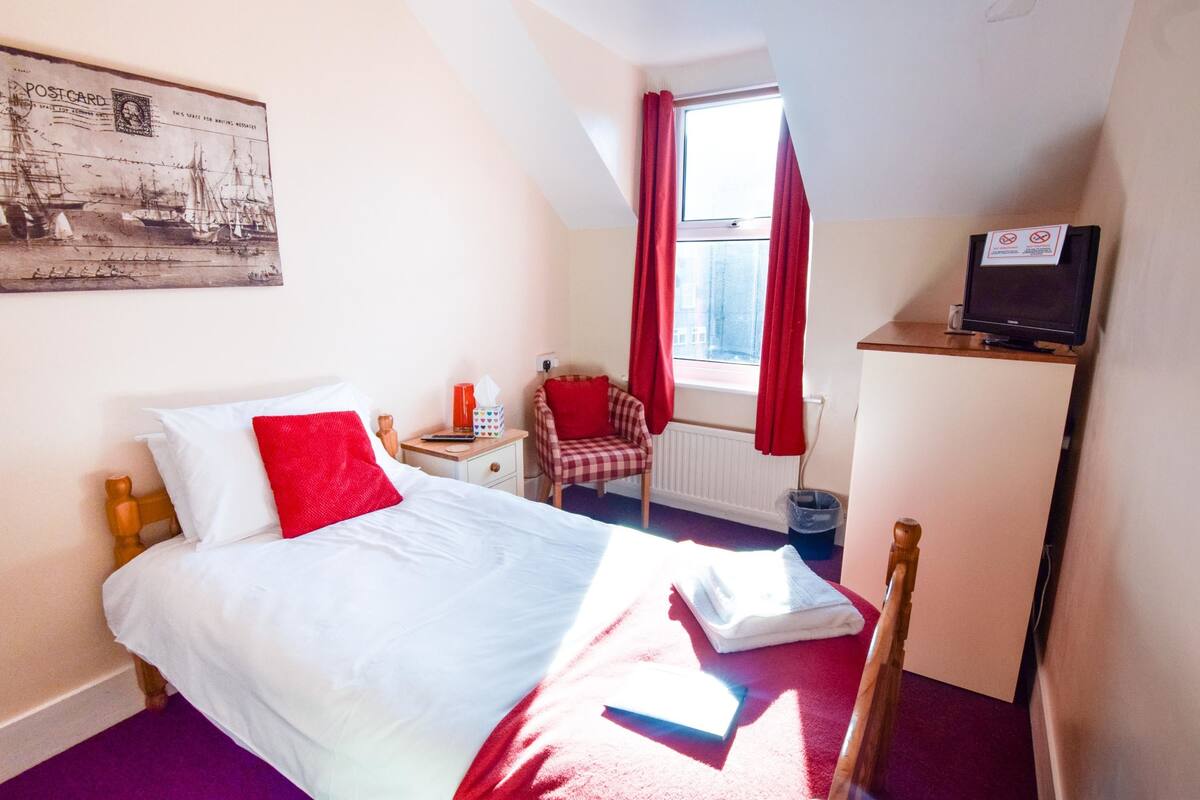 Une chambre d’hôtel avec un lit, une chaise, une télévision et une fenêtre avec des rideaux rouges.