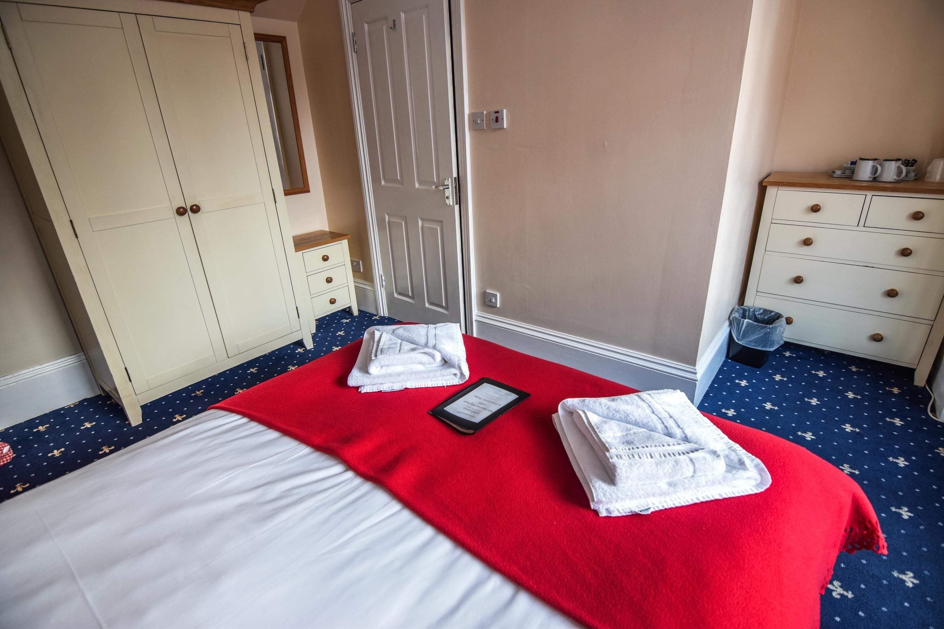 Ein Hotelzimmer mit einem Bett, einer roten Decke, zwei Handtüchern, einem Nachttisch mit Kaffeemaschine und einem Kleiderschrank.