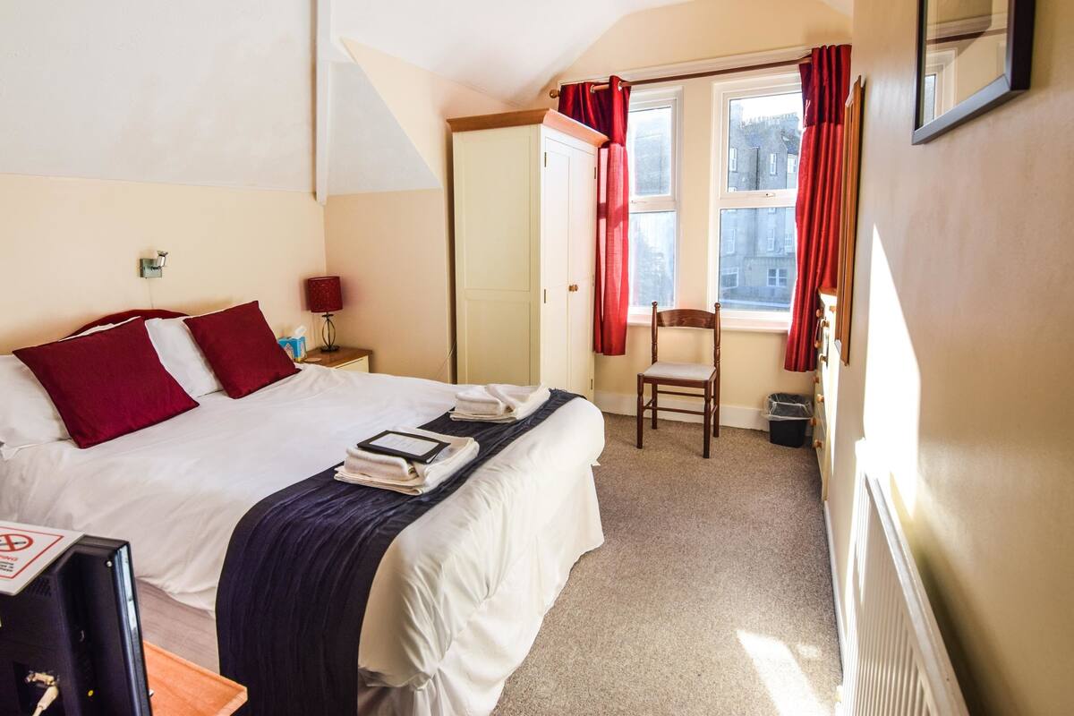 Une chambre d’hôtel avec un lit, une chaise, un bureau, une lampe et une fenêtre avec des rideaux.