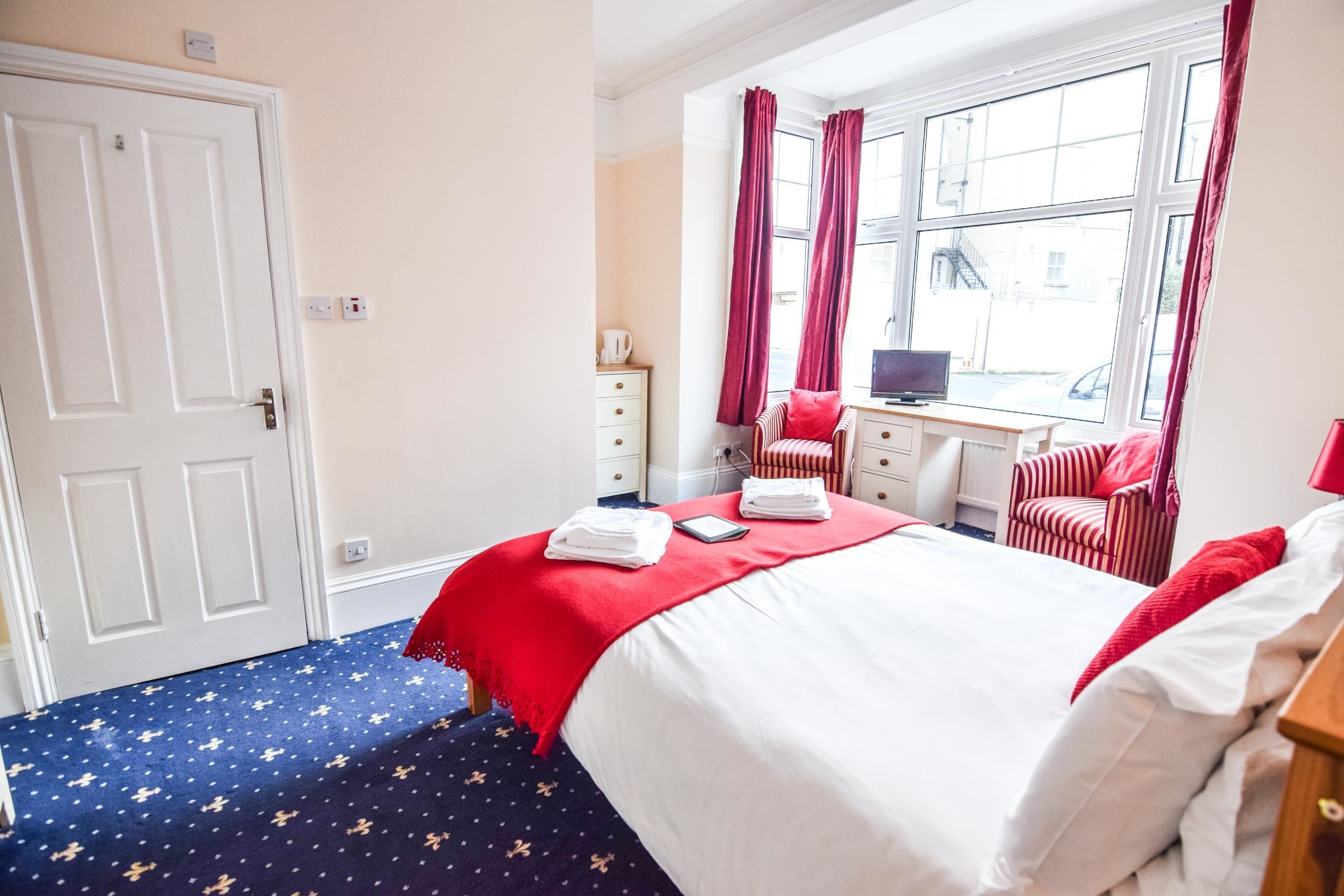 Ein Hotelzimmer mit einem Bett, einem Schreibtisch mit Computer, einem Stuhl und einem Fenster mit Vorhängen.