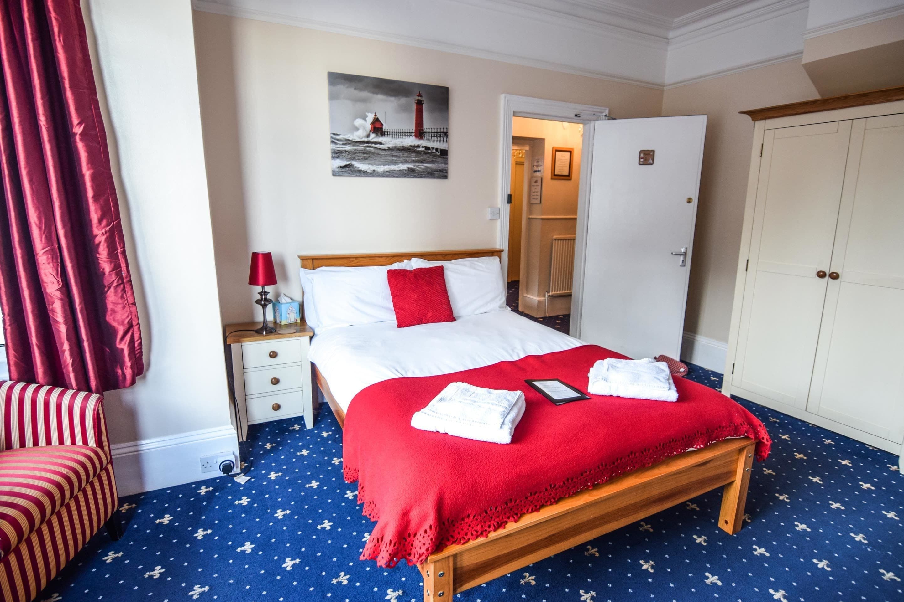 Habitación de hotel ordenada con una cama de madera, ropa de cama roja y blanca, un plaid rojo y una alfombra azul con diseños blancos.