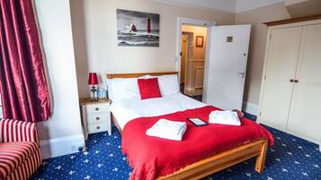 Habitación de hotel ordenada con una cama de madera, ropa de cama roja y blanca, un plaid rojo y una alfombra azul con diseños blancos.