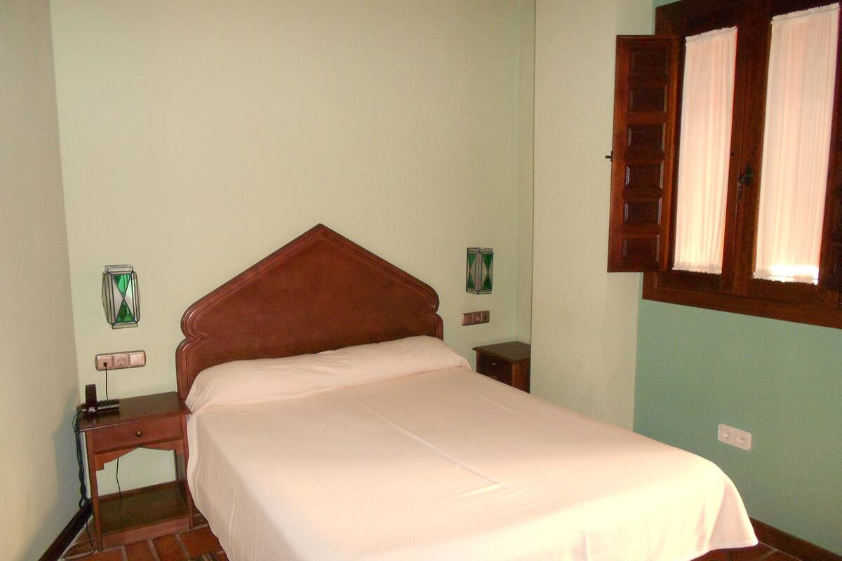 A bedroom with a single bed, a nightstand, a wall socket, and a window with curtains.