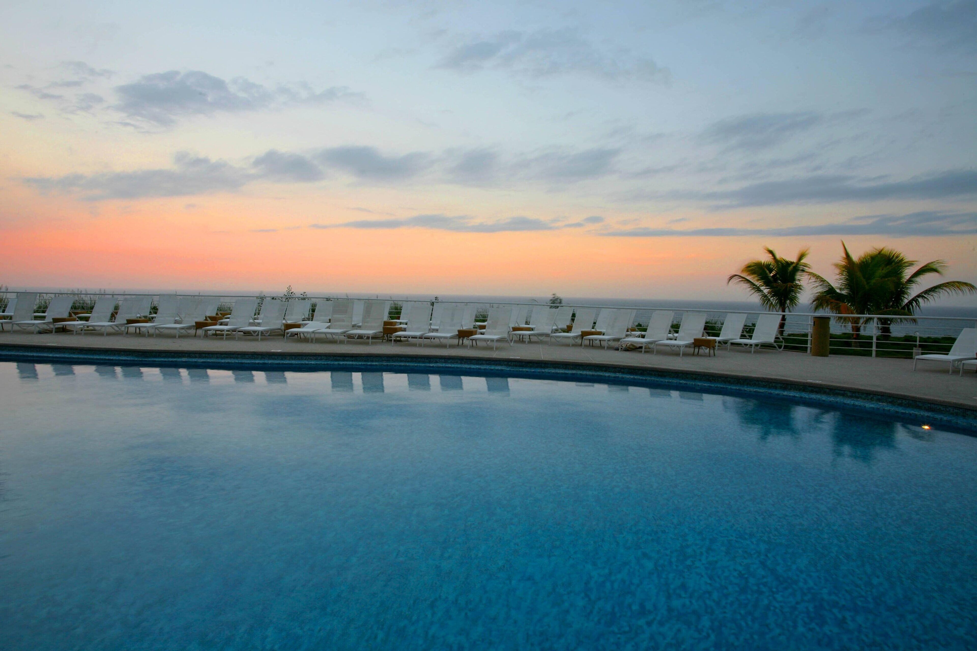 A swimming pool with lounge chairs and a view of the ocean at sunset.