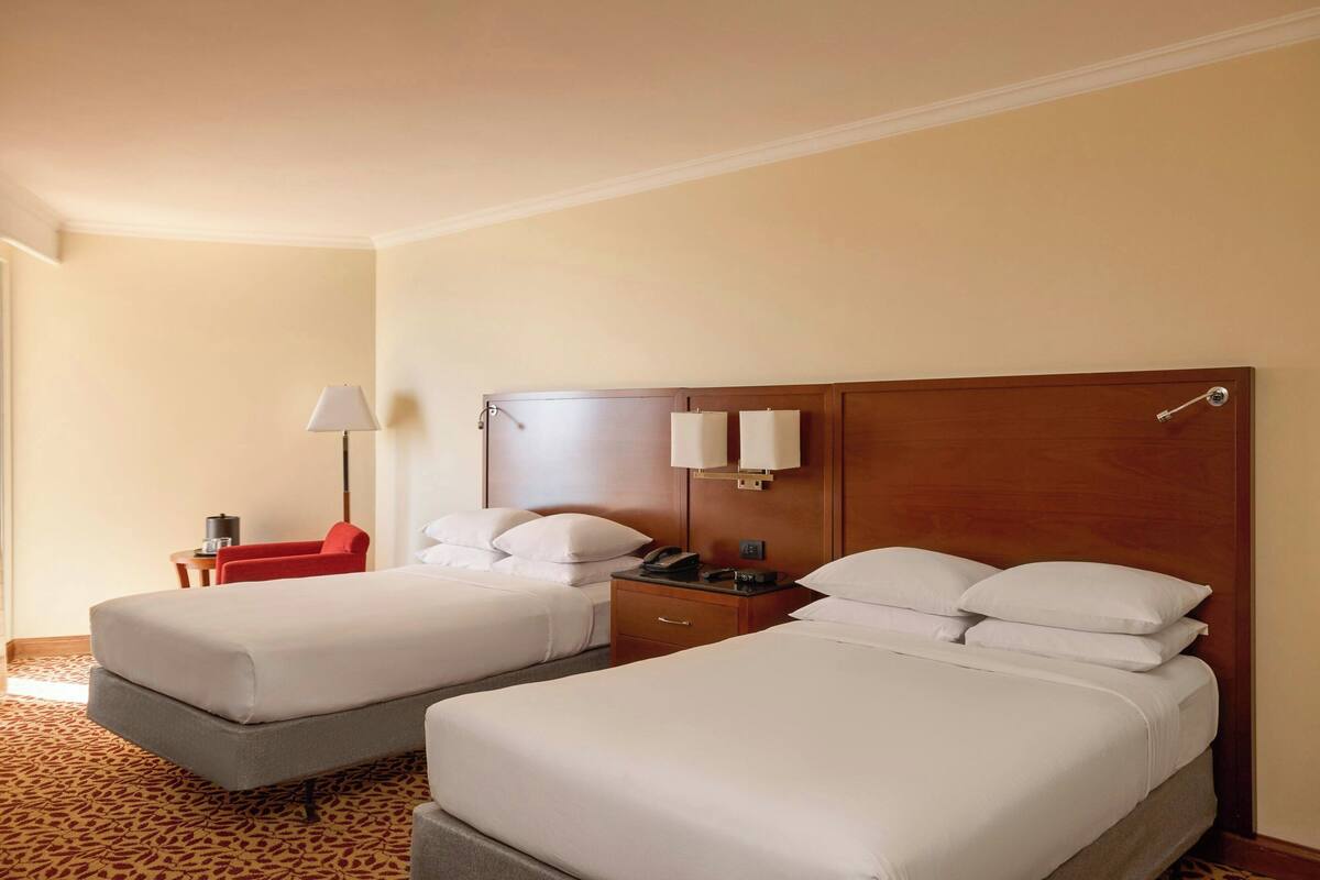 Une chambre d’hôtel avec deux lits, une chaise rouge, une table de chevet avec une lampe et une tête de lit en bois.