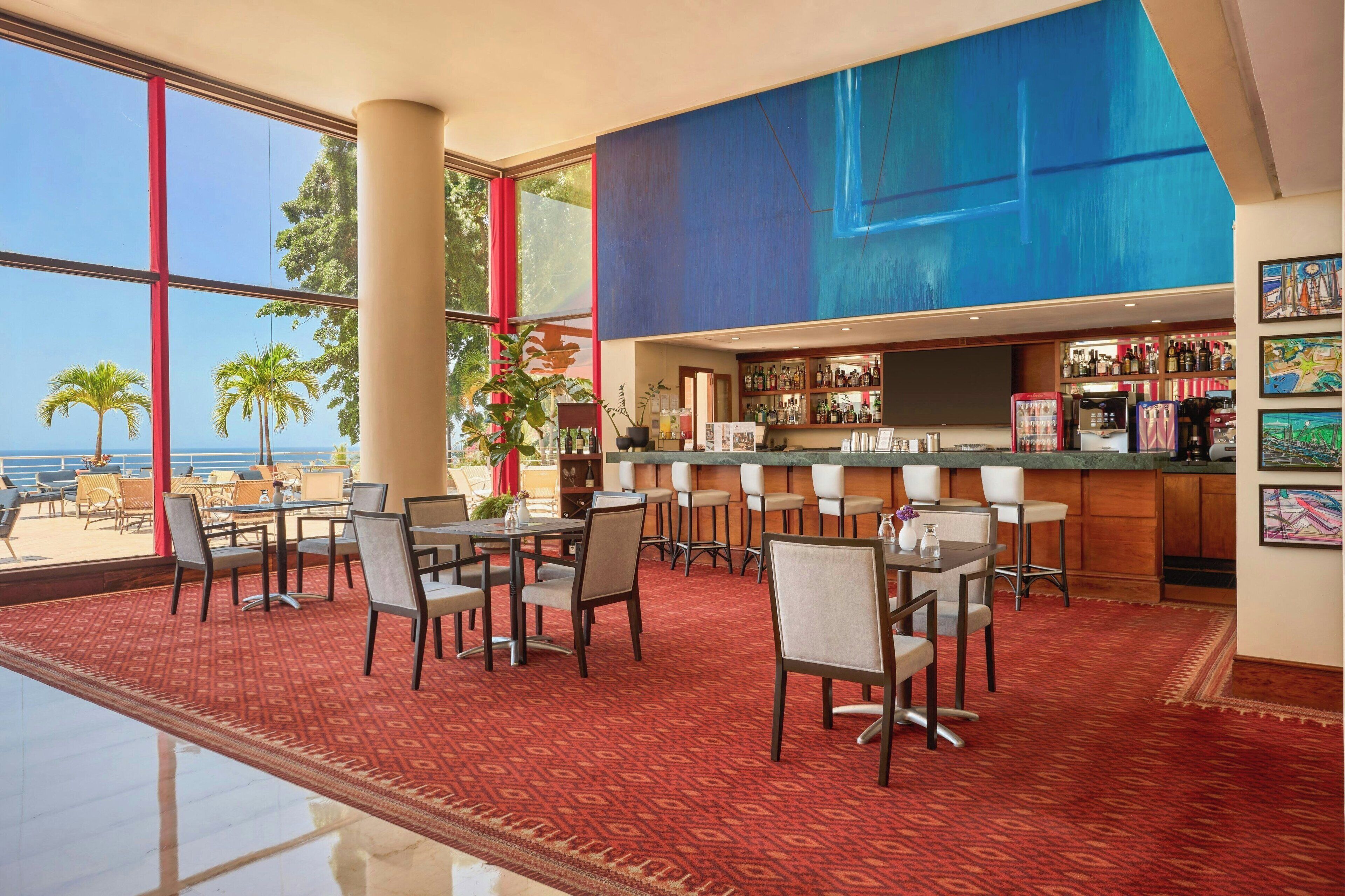A bar area with a view of the ocean, a red carpeted floor, and a bar counter with various bottles and a refrigerator.