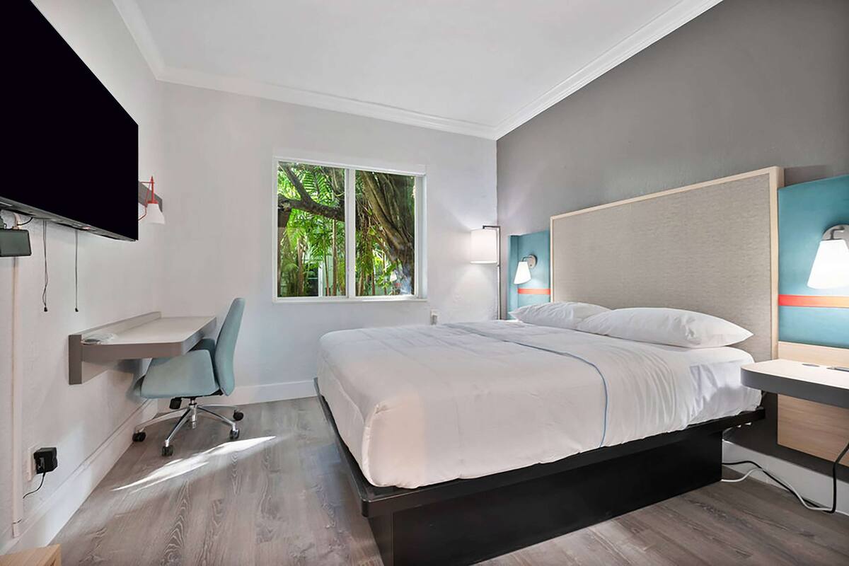 Una camera da letto moderna con un letto, una scrivania, una sedia, una televisione e una finestra con vista sugli alberi.