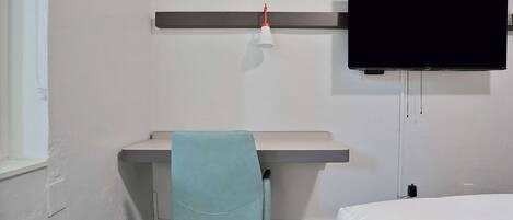 A room with a bed, a desk with a chair, a television mounted on the wall, and a wall-mounted heater.
