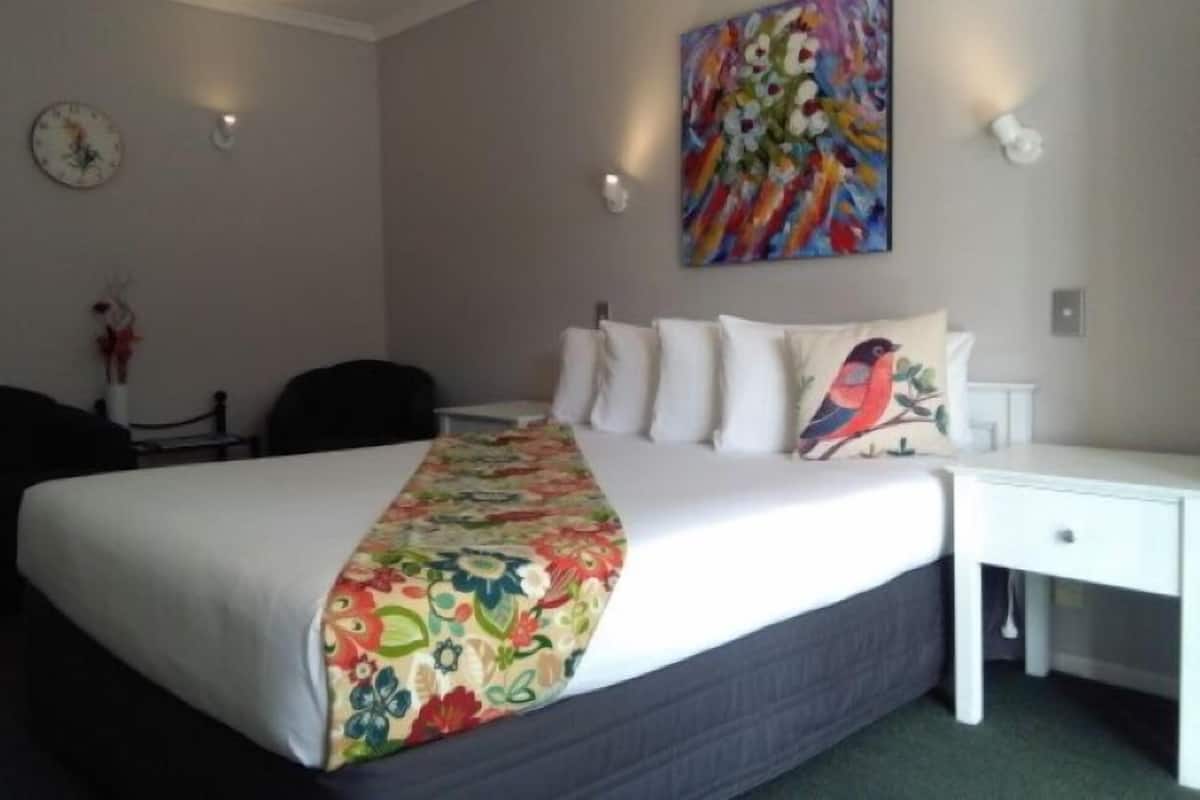 A hotel room with a large bed, a nightstand, a chair, a painting on the wall, and a clock.