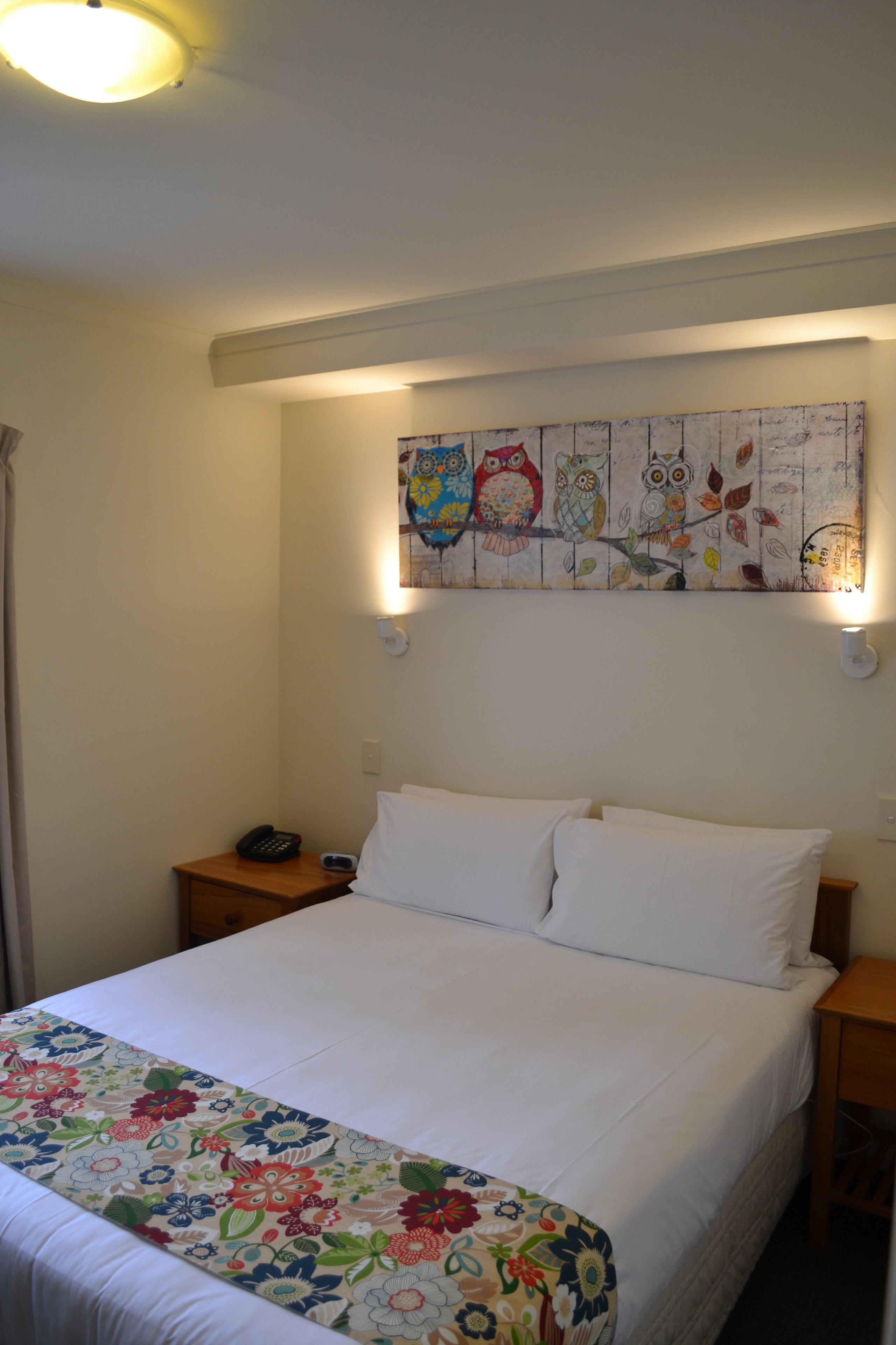 Een keurig opgemaakt bed met een bloemenbedekking, een aan de muur hangend schilderij van uilen en bedlampen.