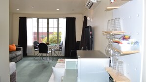 A hotel room with a sofa, a dining table with chairs, a refrigerator, and shelves with glassware. - Colonial Lodge Motel (Taupo)