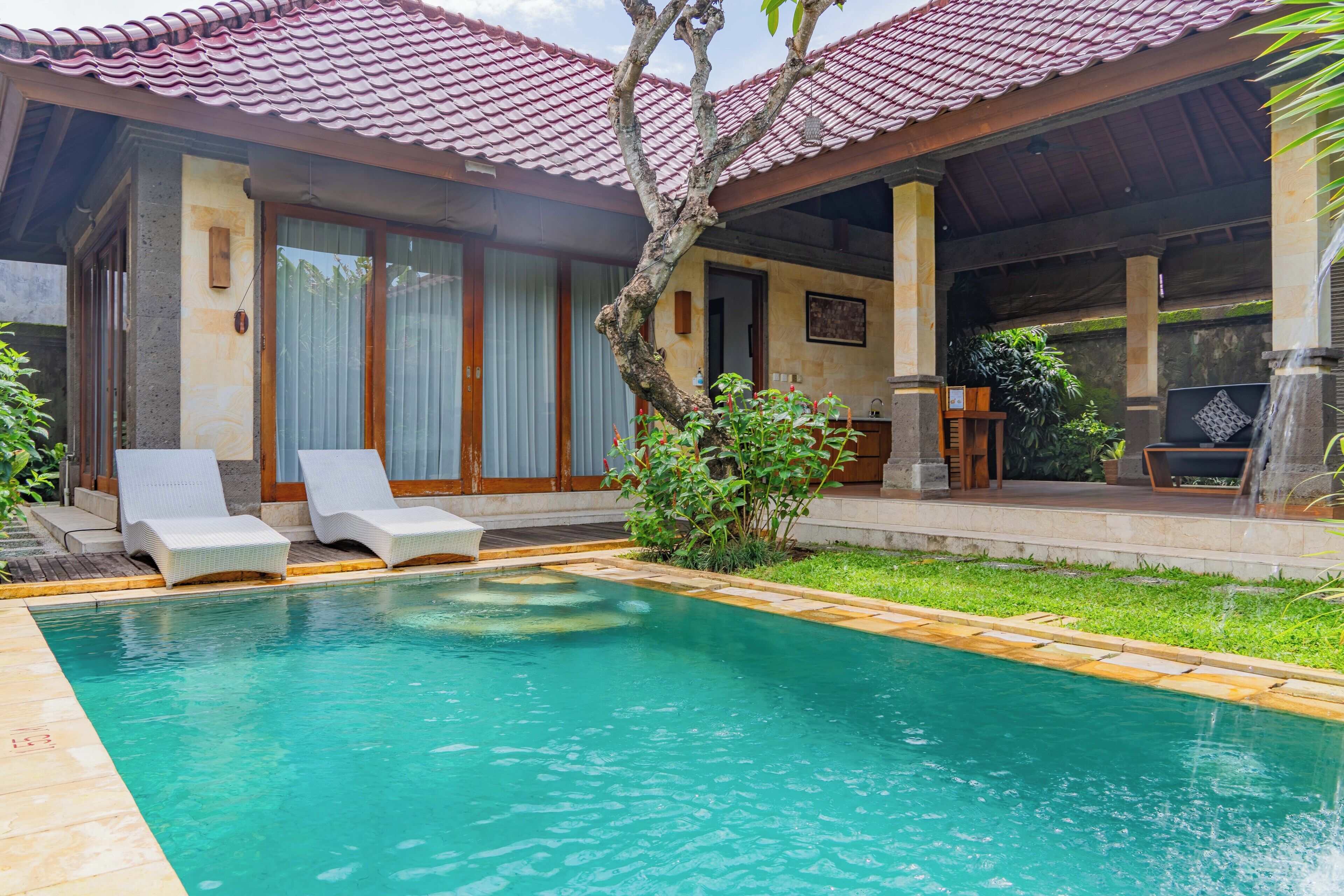 A swimming pool with lounge chairs, a covered patio, and a view of a building with large windows and a tiled roof.