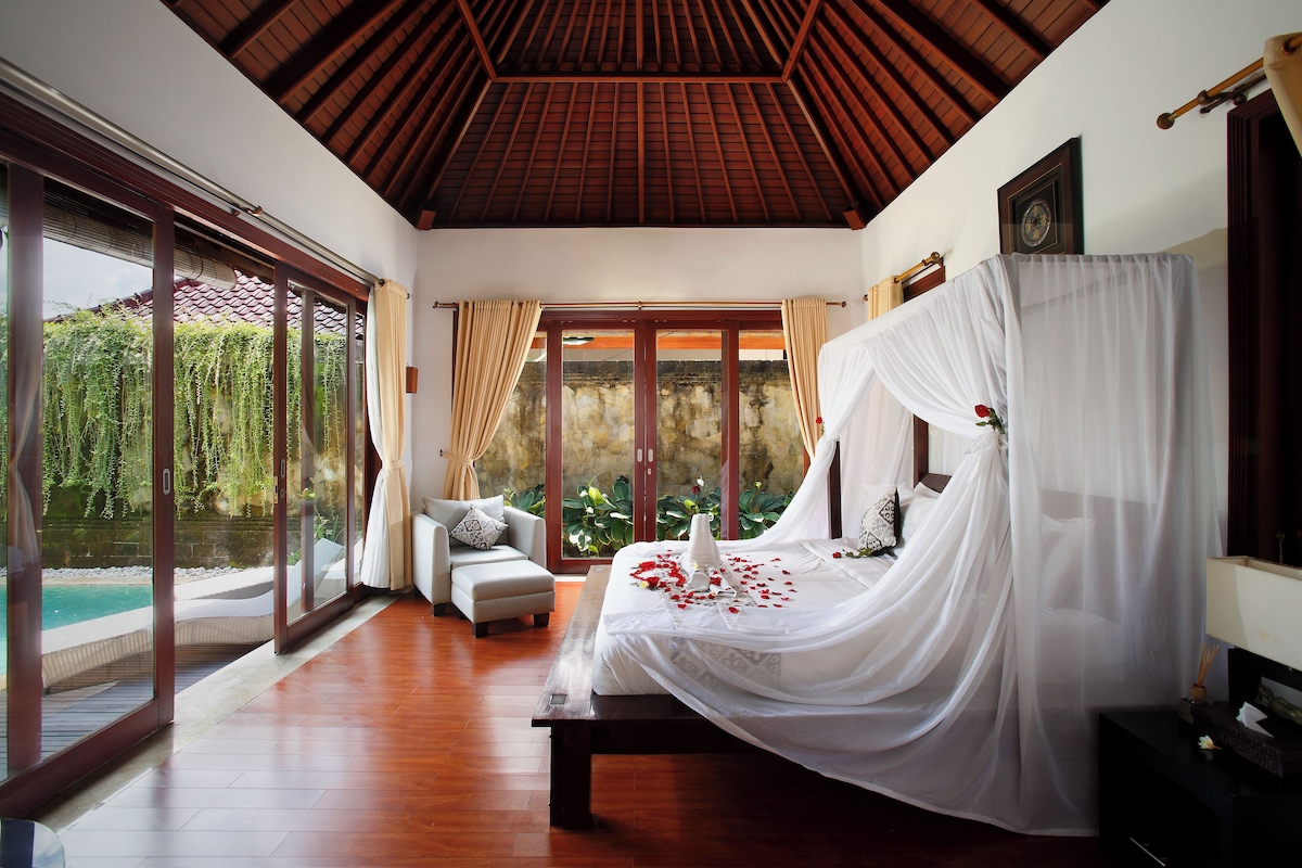 Ein Schlafzimmer mit einem großen Bett, Blick auf einen Pool und einer Holzbalkendecke.
