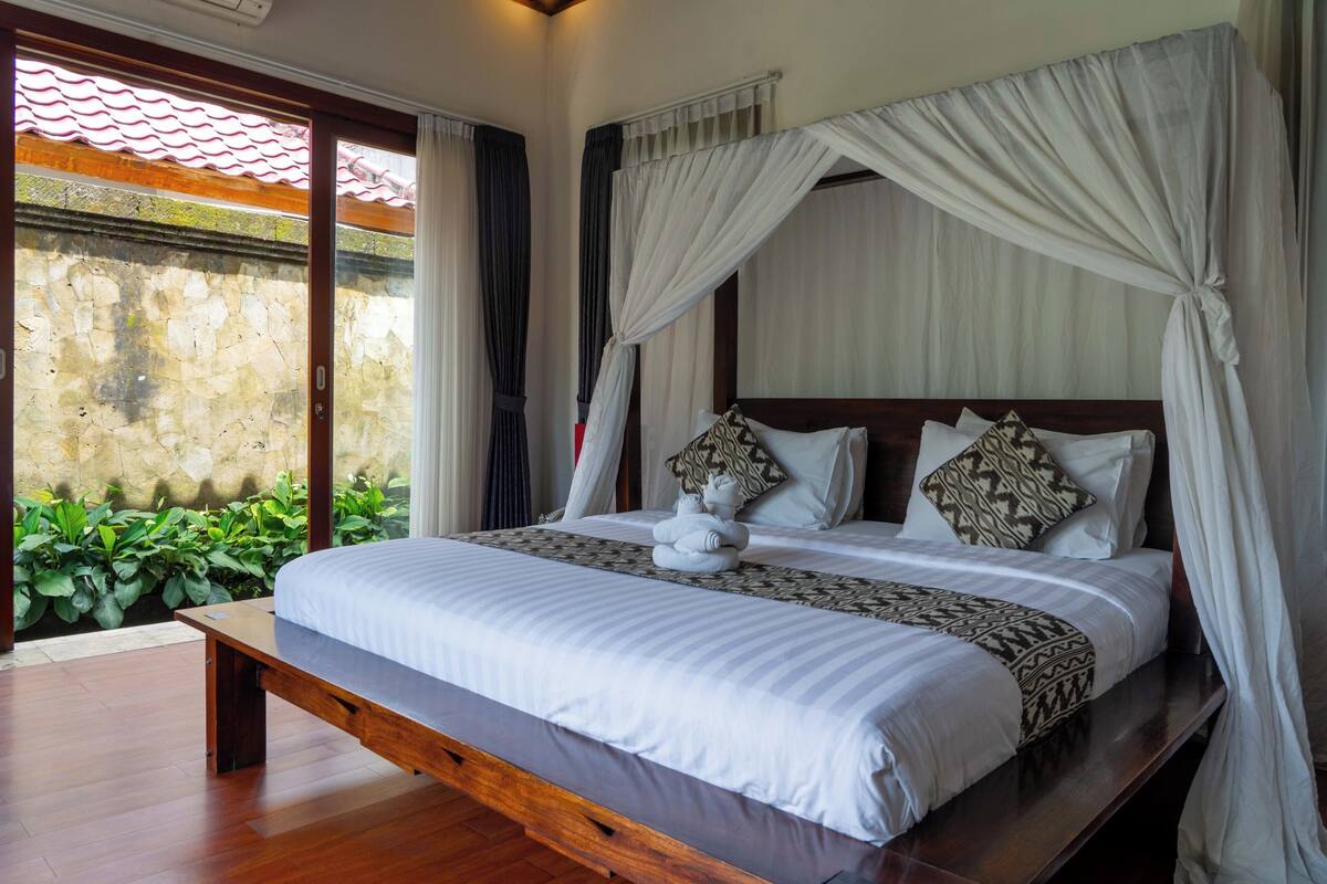 A large bed with a canopy, a bench at the foot of the bed, and a view of greenery outside.