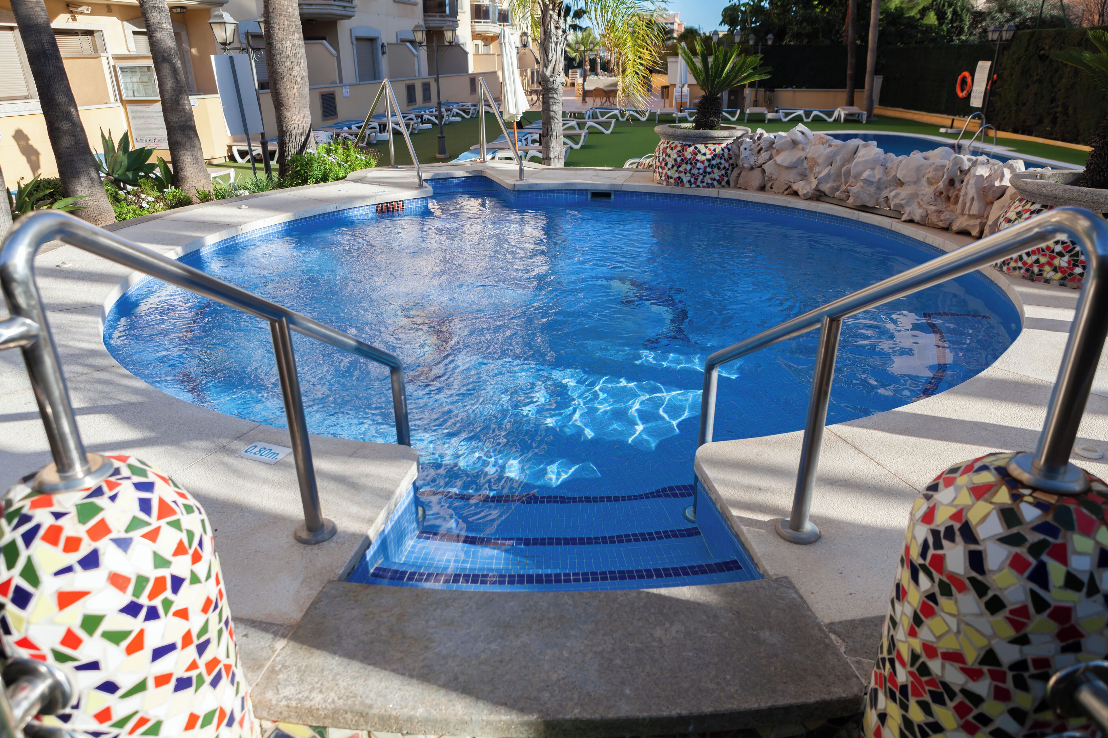 Una piscina con borde de baldosas, barandillas de metal y un muro de piedra que la rodea.