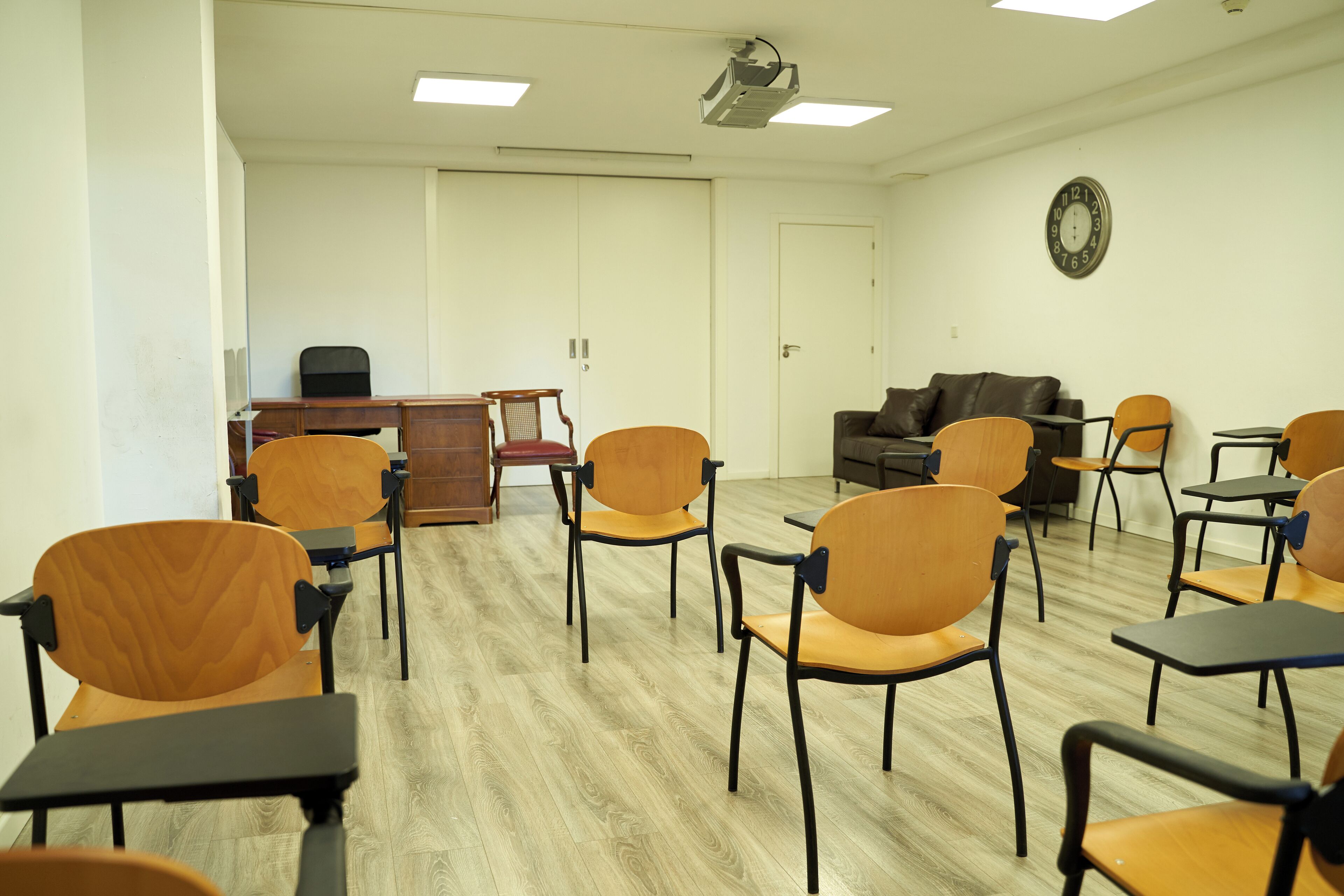 Un aula con sillas y escritorios de madera, un sofá de cuero marrón y un reloj en la pared.