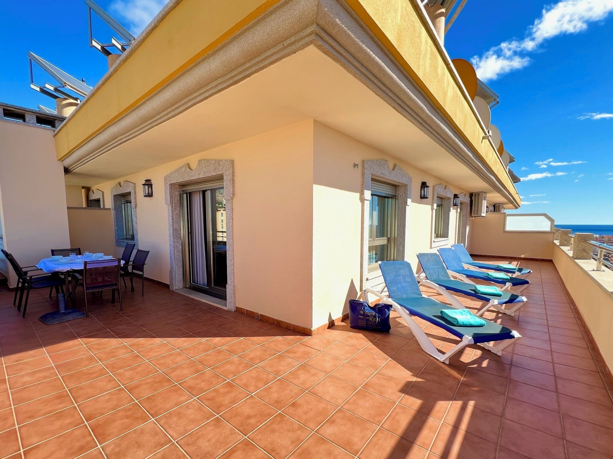 A sunlit balcony with a dining table and chairs, a sliding glass door, and several sun loungers.