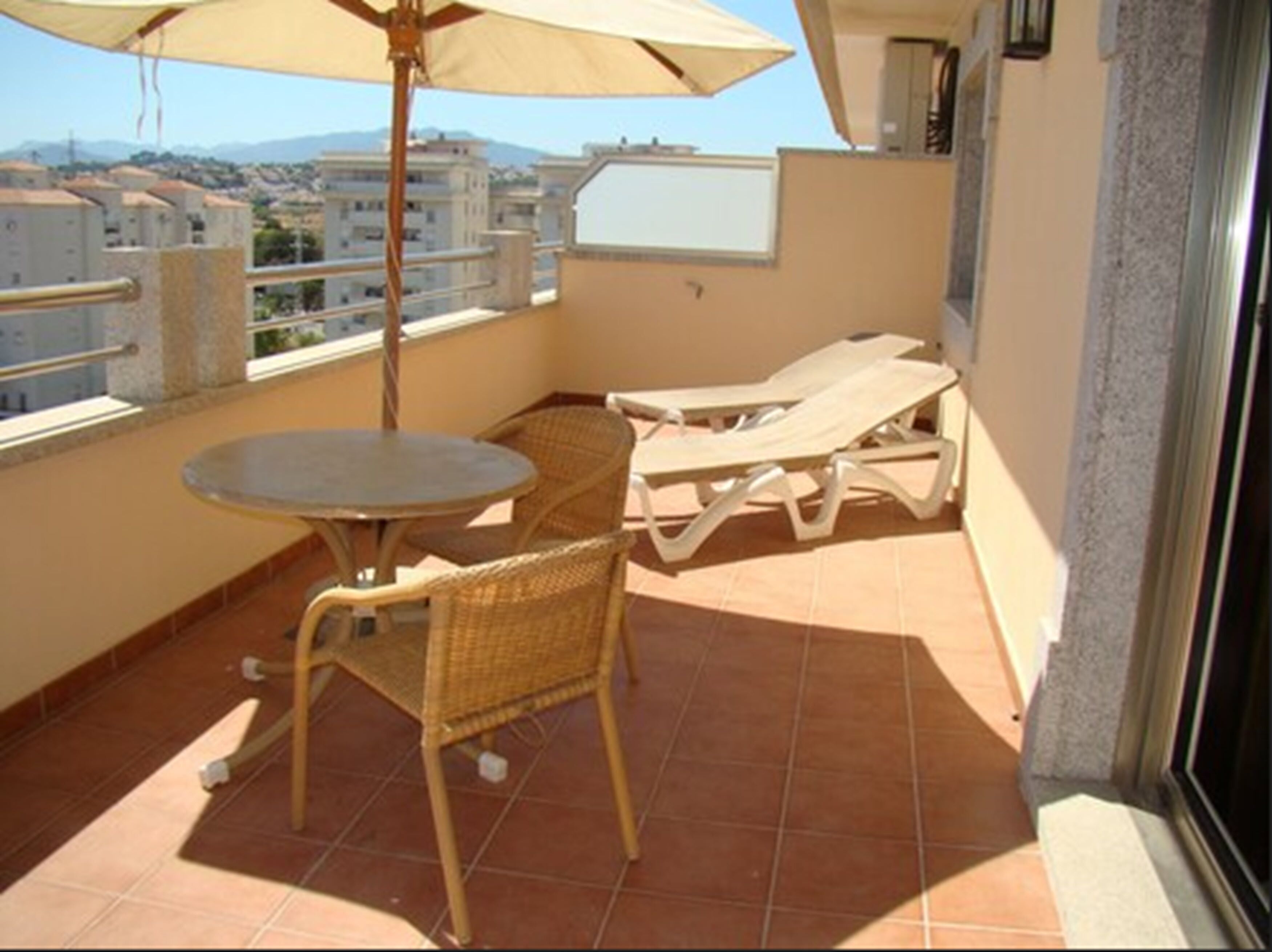 Un balcón con una mesa redonda y dos sillones, una sombrilla y vistas a edificios al fondo.