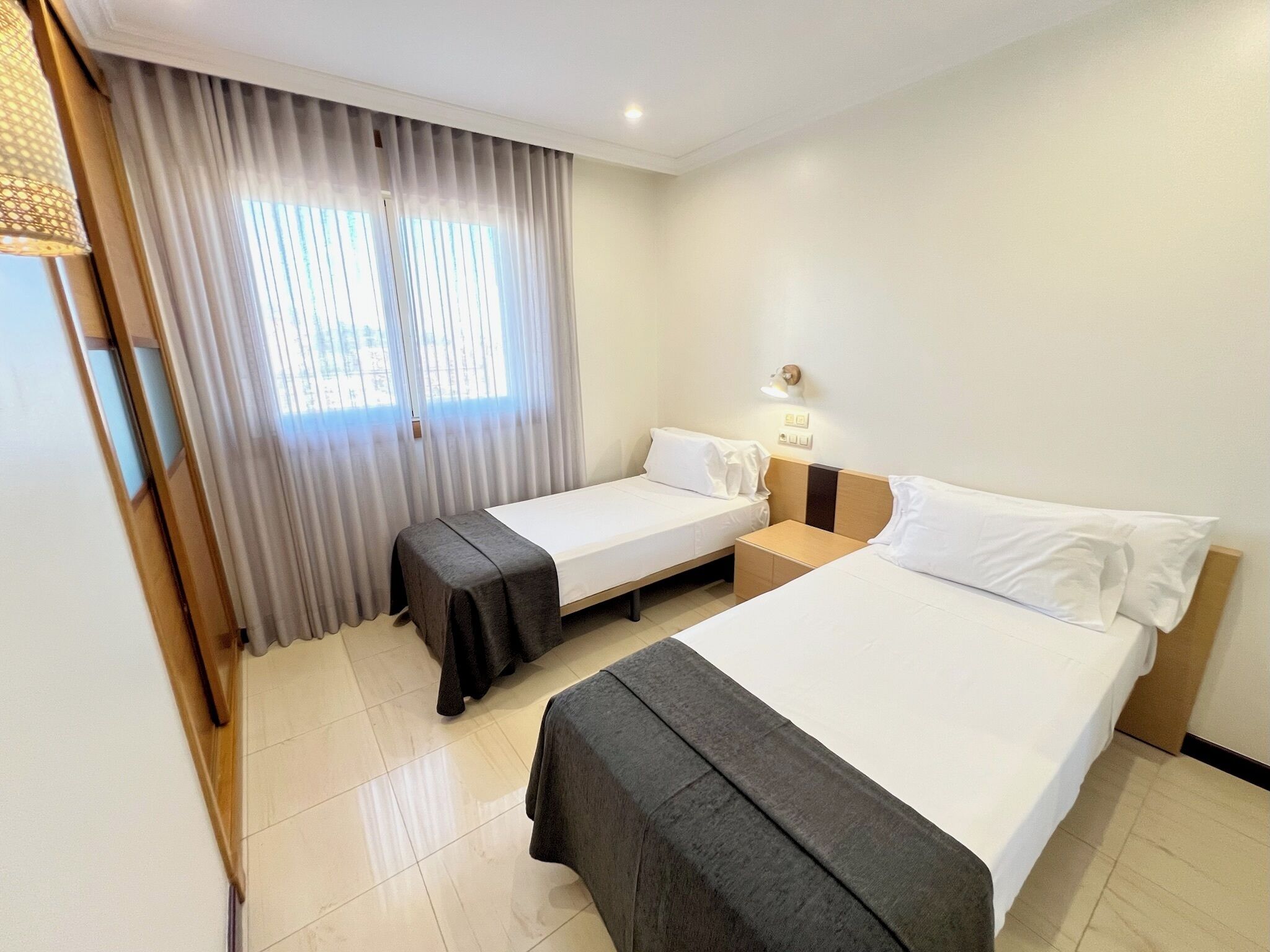 A hotel room with two beds, a nightstand, a lamp, and a window with curtains.