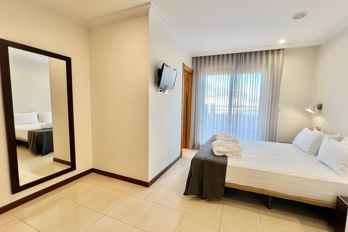 A hotel room with a large bed, a flat-screen TV mounted on the wall, a bedside table with a lamp, and a window with curtains.