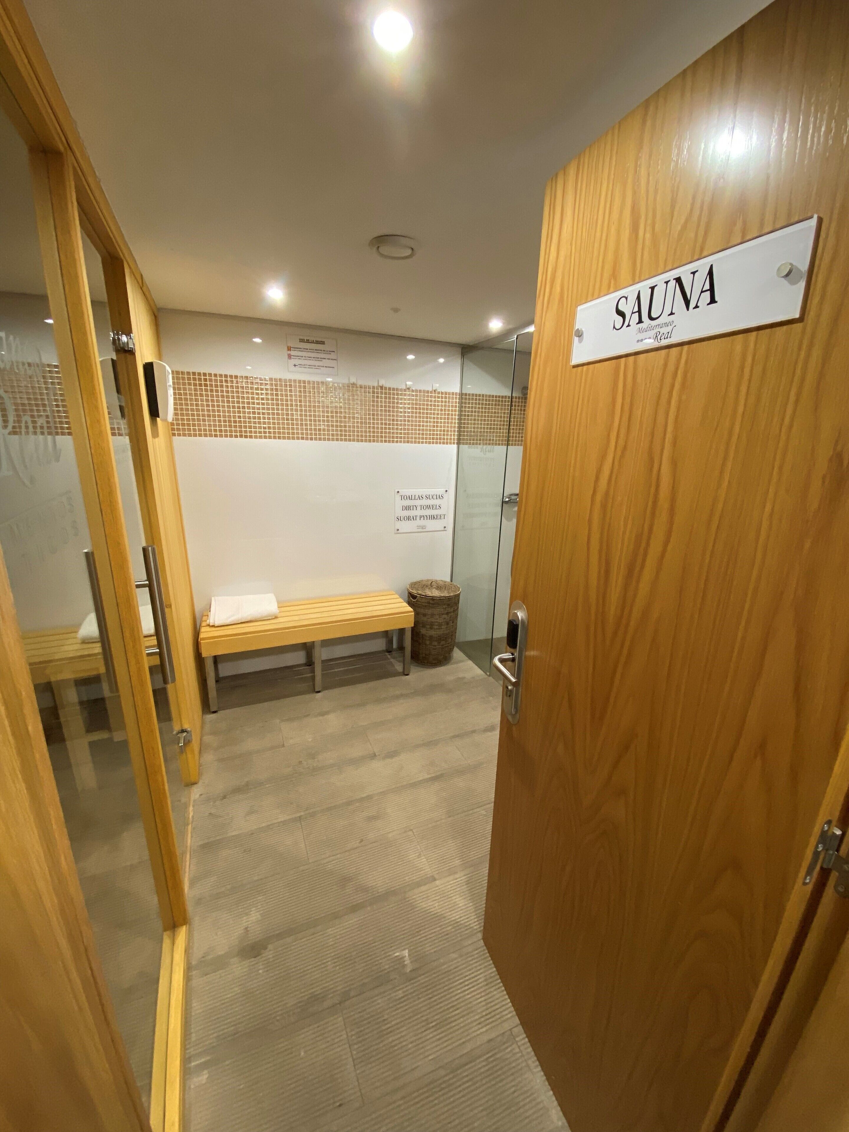 A sauna room with a wooden door labeled "SAUNA," a bench, a trash bin, and a sign instructing to use dirty towels.