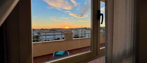 Ein Fenster mit Blick auf eine Skyline bei Sonnenuntergang, ein Tisch mit einem Buch und ein teilweise zurückgezogener Vorhang.