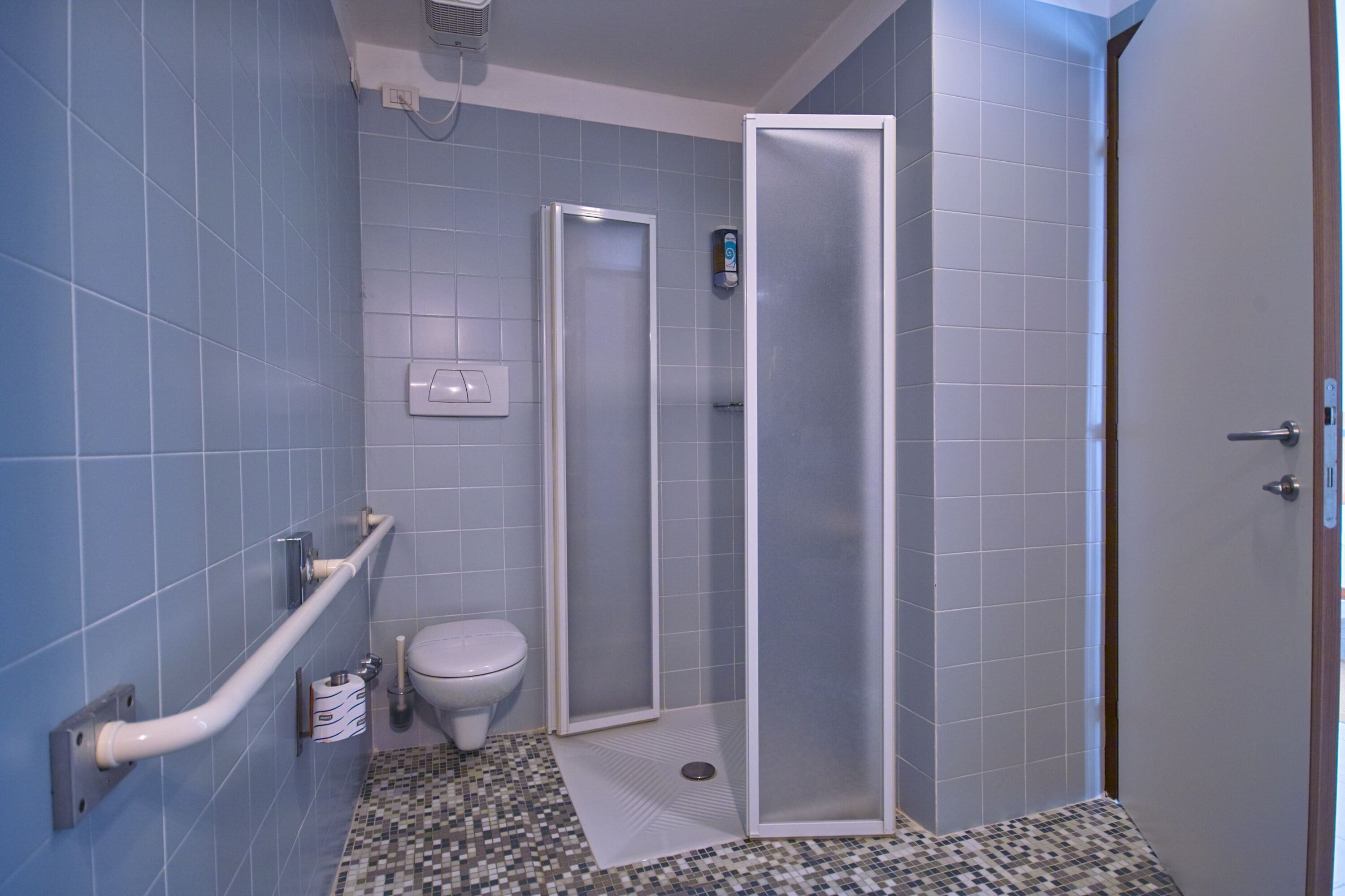 Une salle de bain avec des carreaux bleus, des toilettes, une douche avec une porte en verre et une barre d’appui.