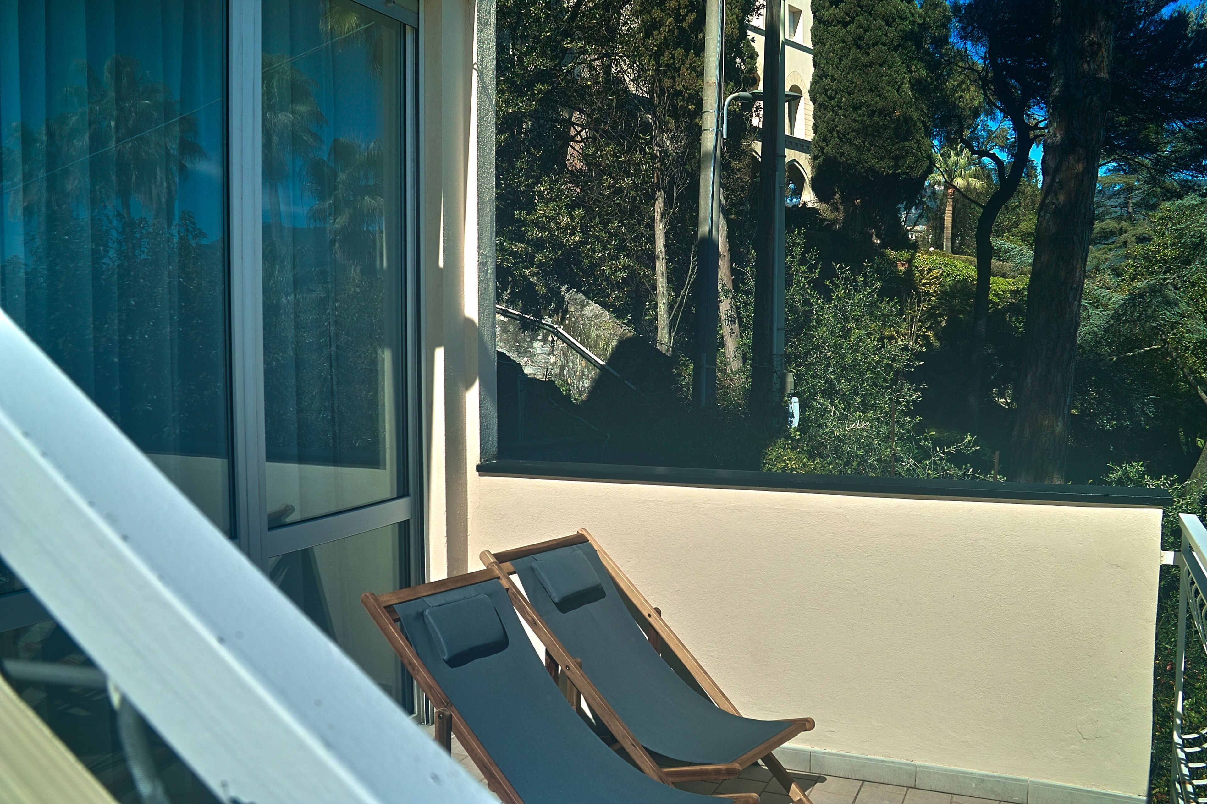 A balcony with two wooden lounge chairs, a glass door, and a view of a lush garden with trees and a clear sky.