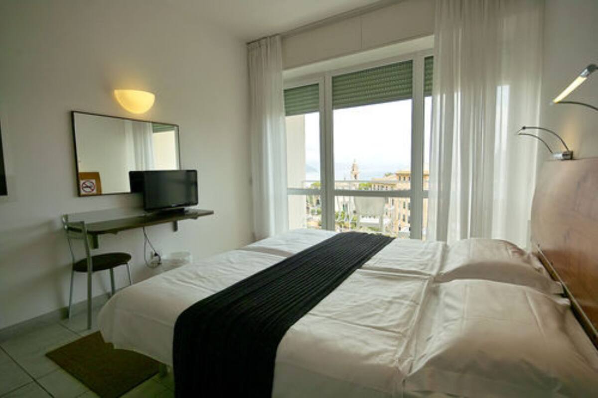 Ein Hotelzimmer mit Bett, Schreibtisch mit Fernseher, Stuhl, Fenster mit Vorhängen und Blick auf Gebäude.