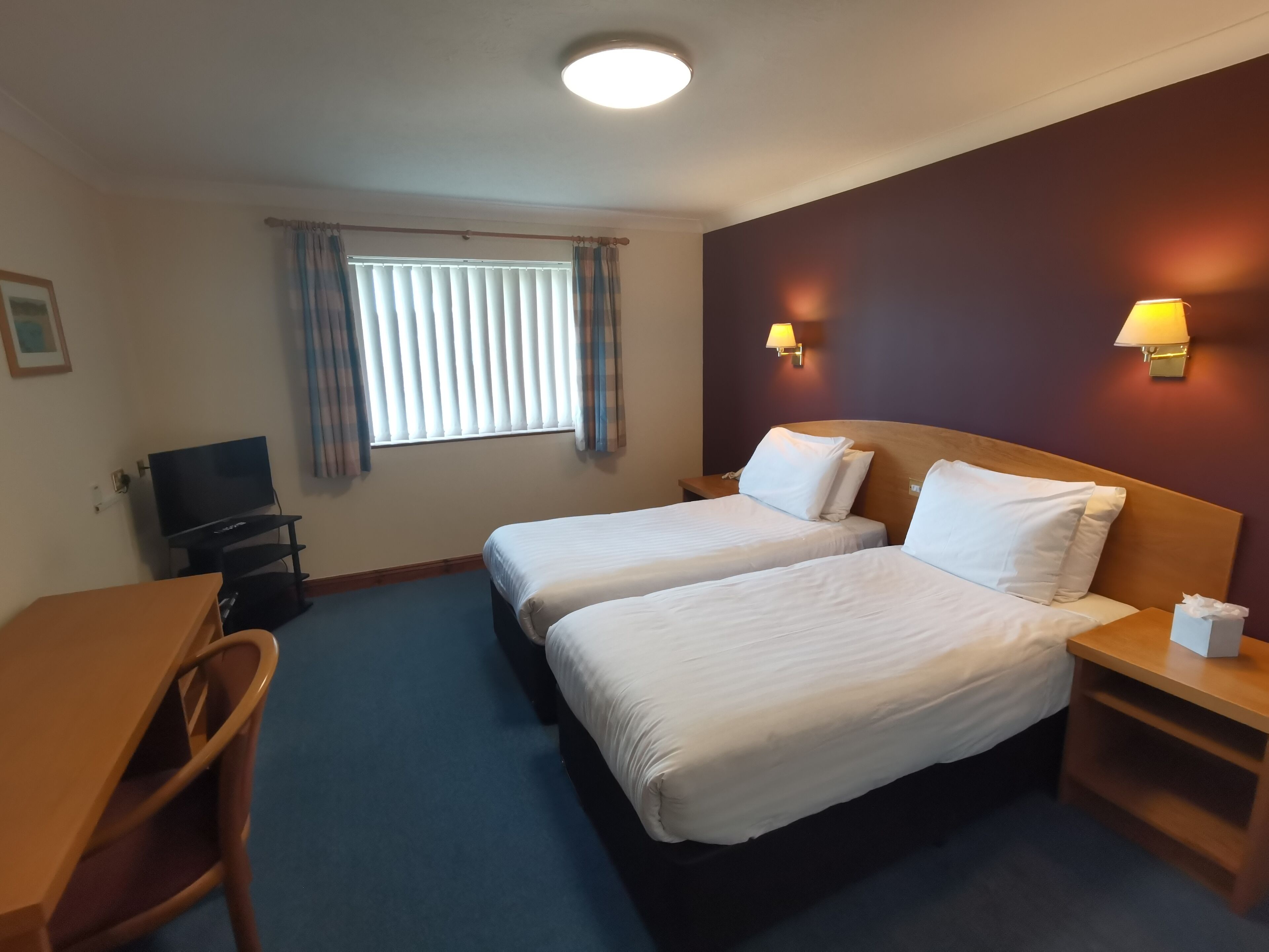 A hotel room with two beds, a desk, a TV, and a window with curtains.