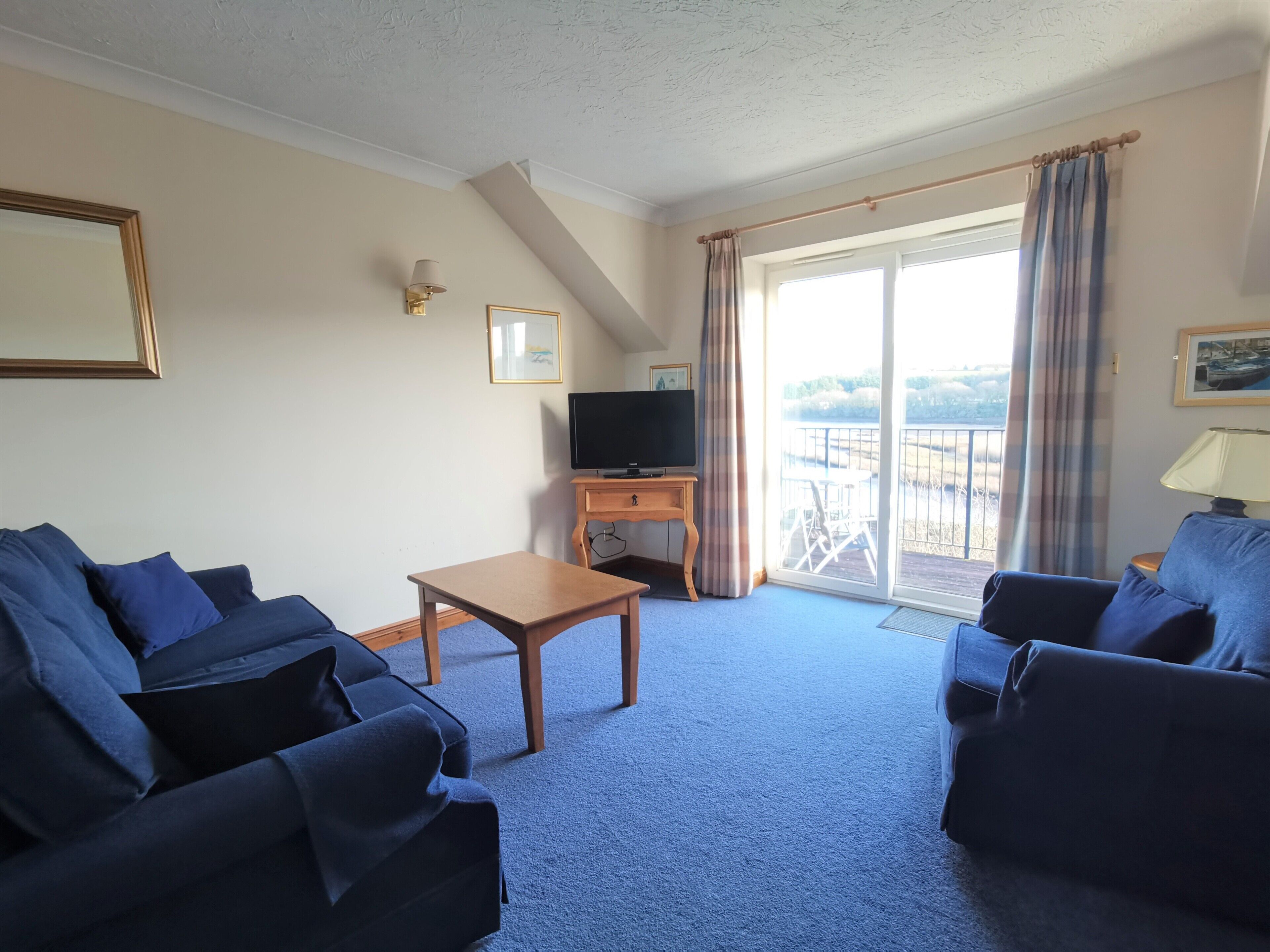 Une chambre d’hôtel avec deux canapés bleu foncé, une table en bois, un téléviseur posé sur un meuble, un balcon avec vue et un tableau encadré au mur.