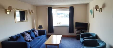 A living room with a sofa, armchairs, a coffee table, a TV, and a window with curtains.