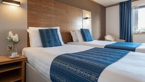 A hotel room with two beds, a wooden headboard, bedside lamps, a chair, a window with curtains, and a small table with a vase of flowers.