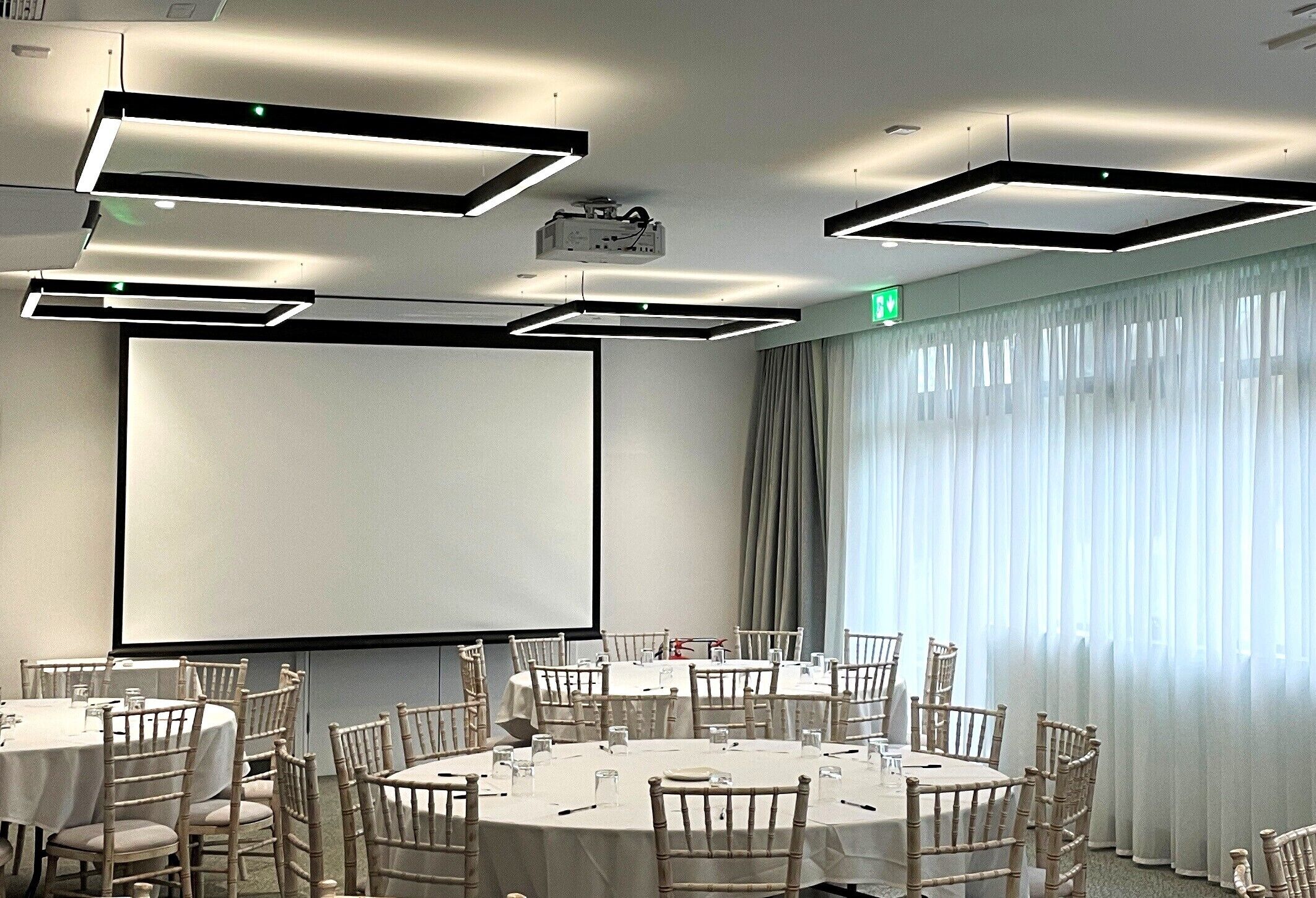 Un salón de conferencias con mesas redondas, sillas, una pantalla de proyector y un amplio ventanal con cortinas.