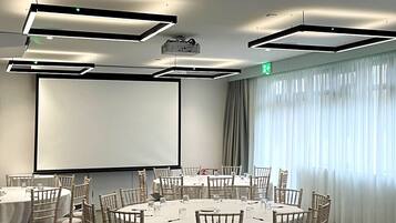A conference room with round tables, chairs, a projector screen, and a large window with curtains.