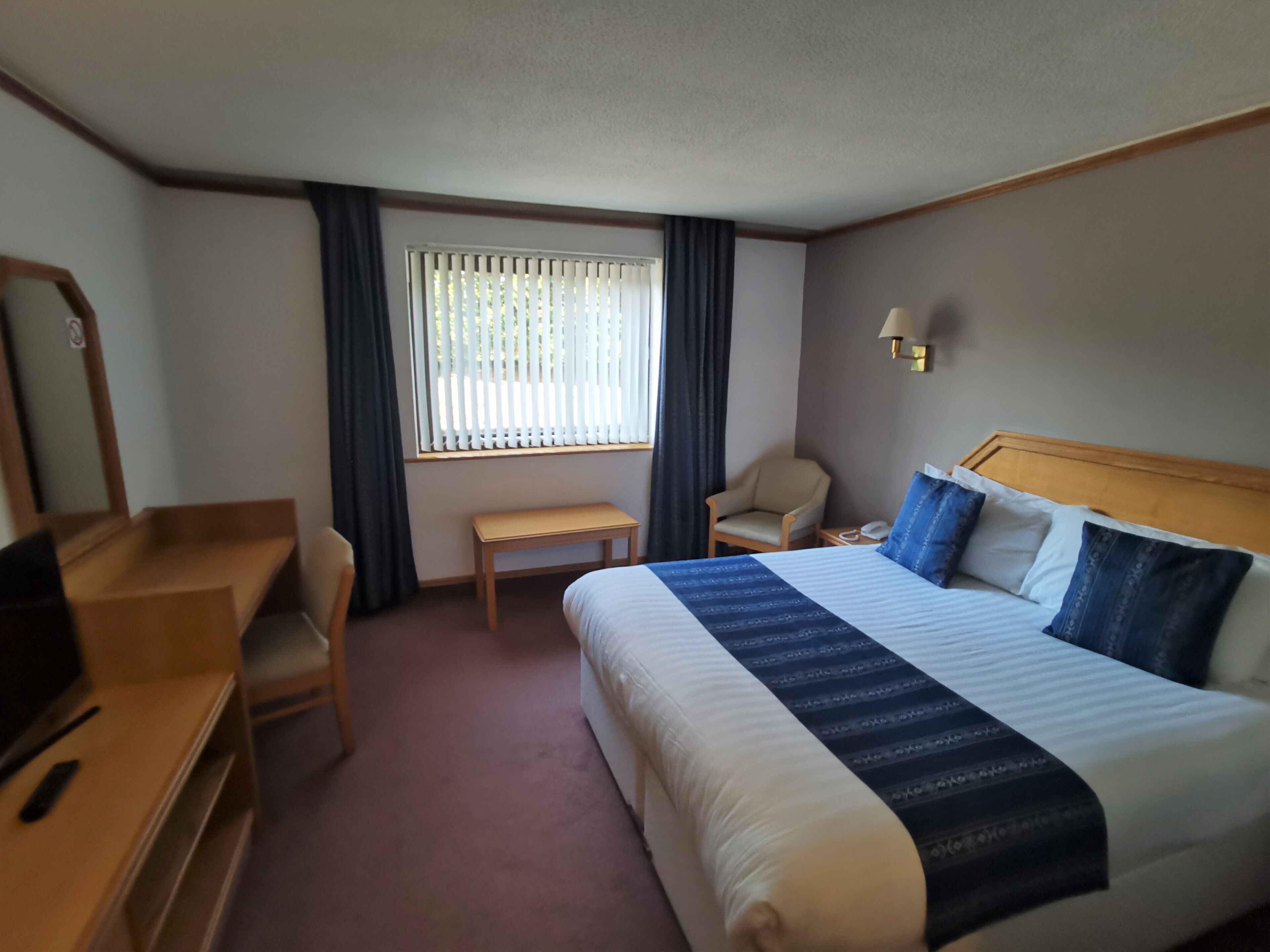 A hotel room with a bed, a desk, a chair, a small table, and a window with curtains.