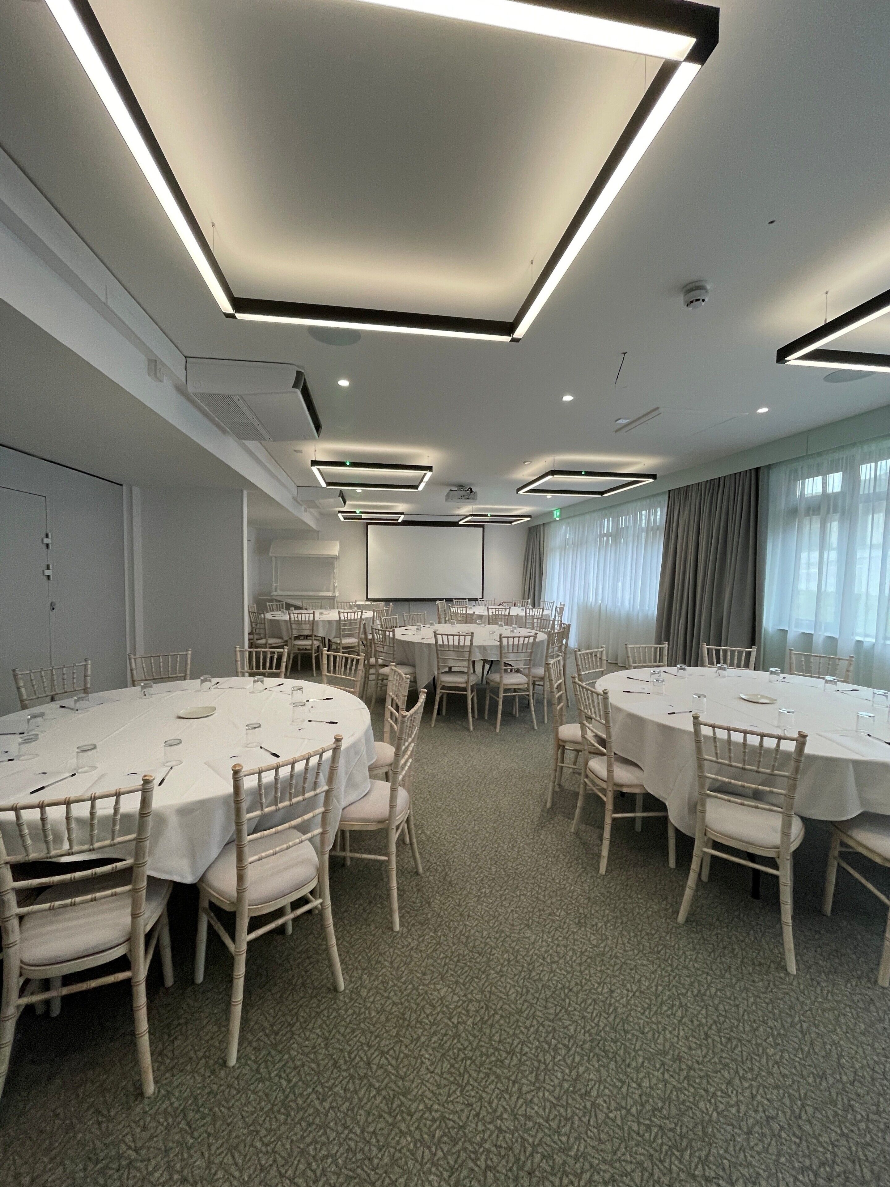 Sala de conferência com várias mesas redondas, cadeiras brancas e luminárias no teto.