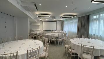 Sala de conferência com várias mesas redondas, cadeiras brancas e luminárias no teto.