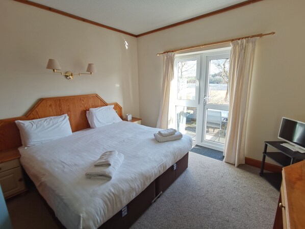 A hotel room with a large bed, a desk with a computer, and a sliding glass door to the outside.