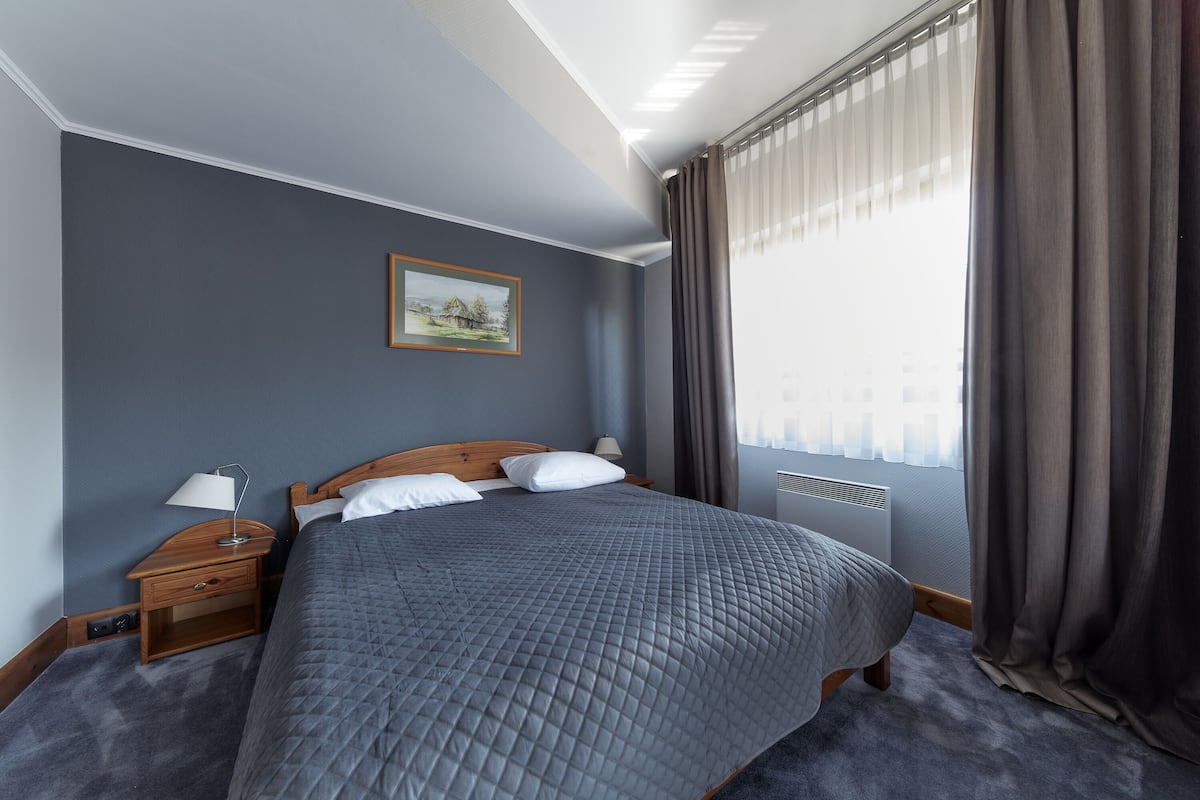 Un dormitorio ordenado con una cama grande, una mesita de noche con lámpara, un cuadro en la pared y una ventana con cortinas.