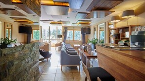 A modern hotel lobby with a stone reception desk, comfortable seating, a kitchen area, and large windows offering a view of the outdoors. - Skalny (Zakopane)