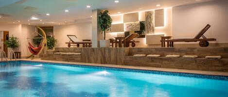 Una piscina interior con sillones de descanso, una hamaca y una pared de piedra con una planta.