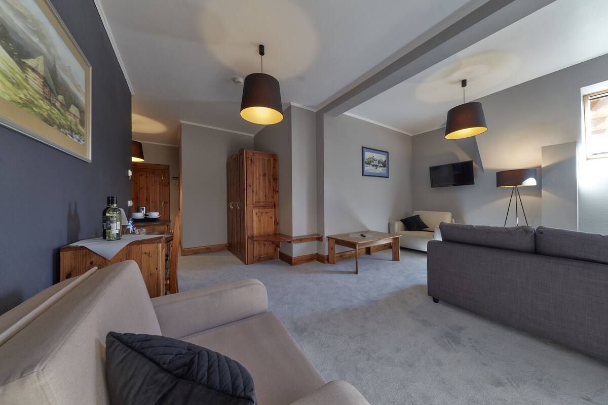 Una sala moderna con un sofá gris, un sillón color beige y un aparador de madera. Hay un televisor de pantalla plana montado en la pared y una lámpara de pie.