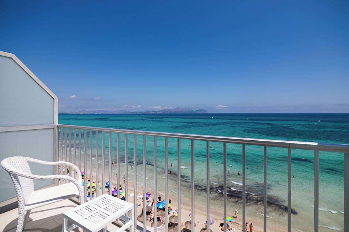 Varanda com cadeiras de vime branco e uma mesa, com vista para uma praia de águas turquesa cristalinas e pessoas nadando.
